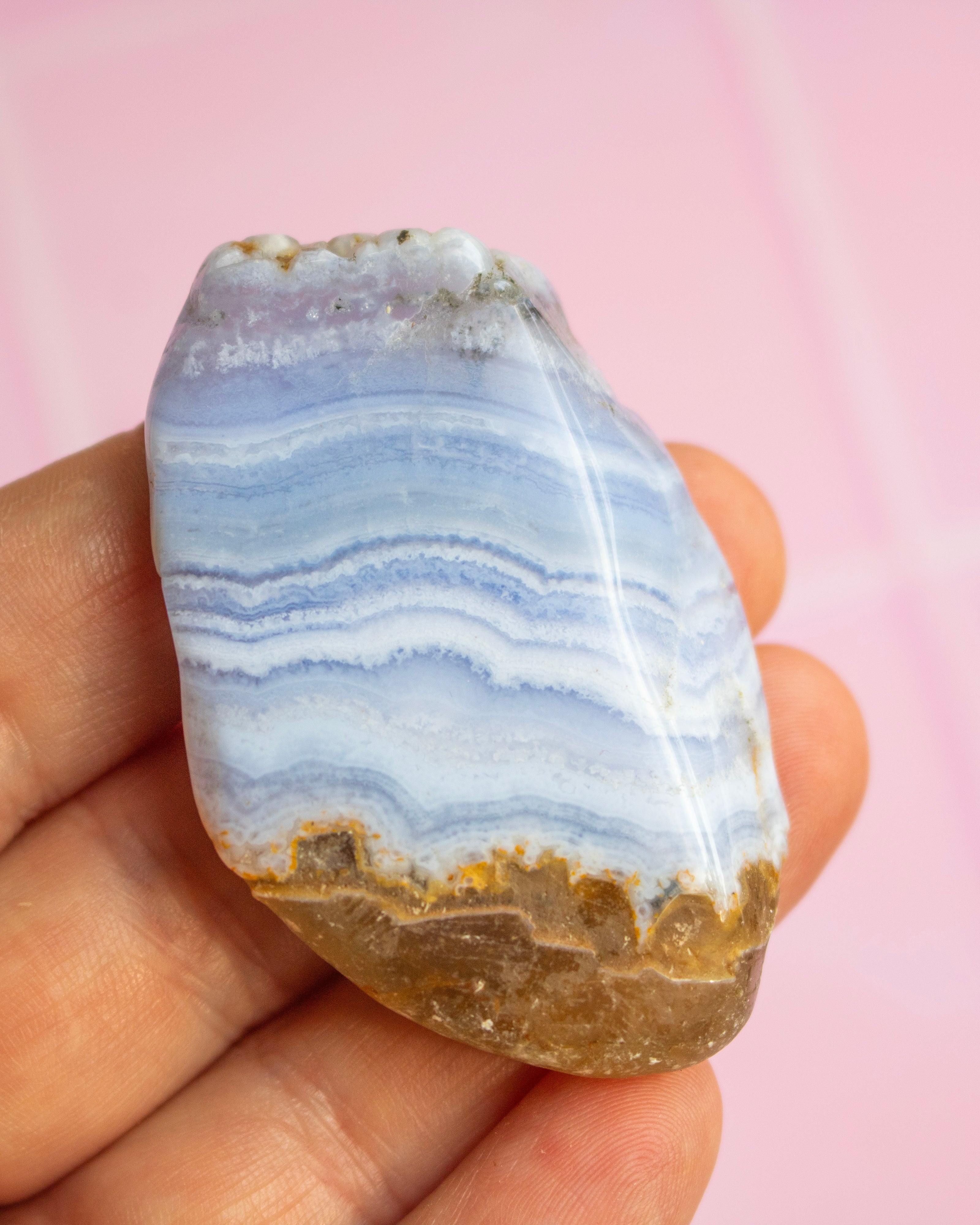 A polished stone with layered blue, white, and gray bands and a brown crystalline base, held in a person's hand against a soft pink background.