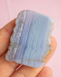 A hand holding a polished rectangular stone with natural wavy banding in pale lavender, light blue, and white, against a soft pink background.