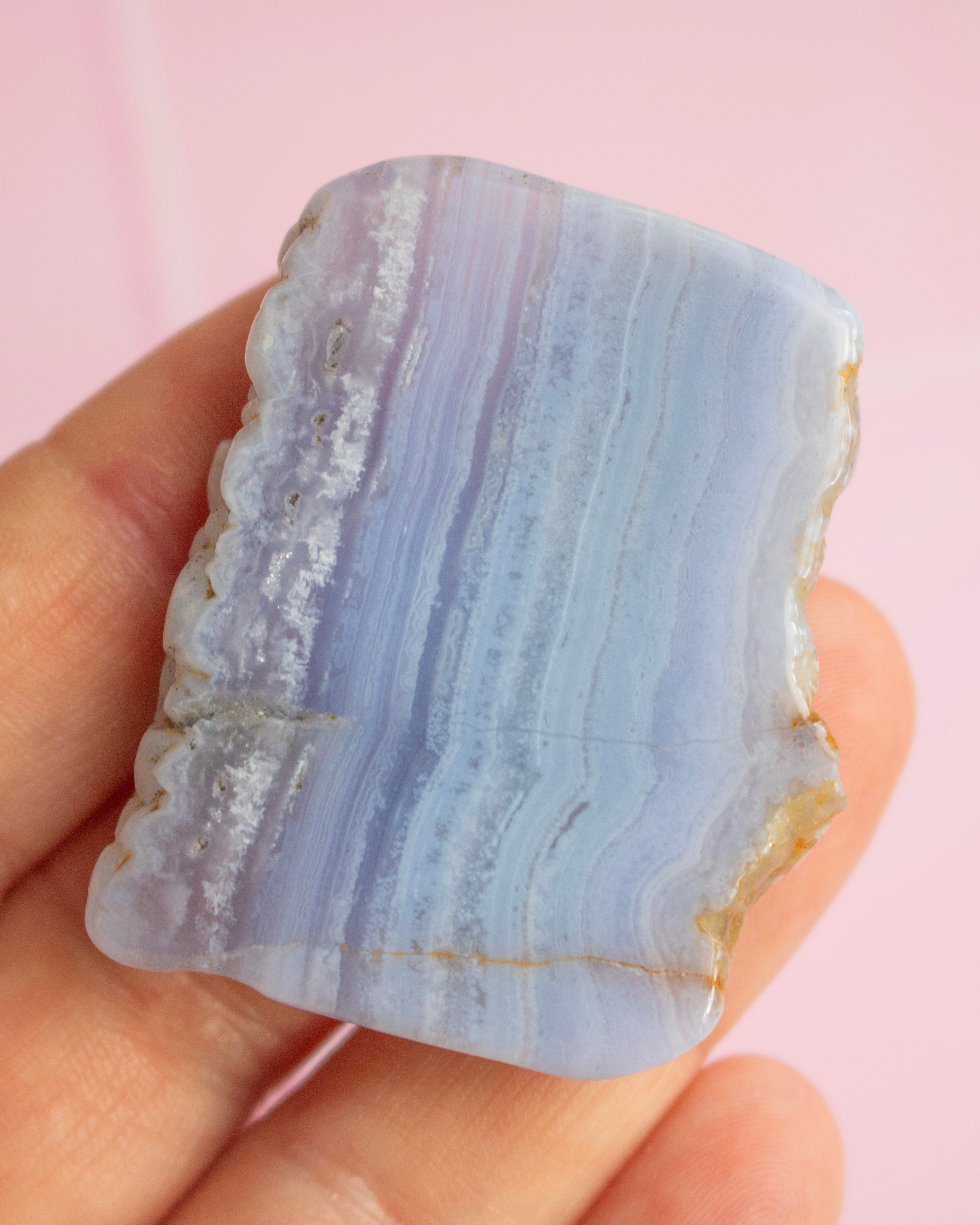 A hand holding a polished rectangular stone with natural wavy banding in pale lavender, light blue, and white, against a soft pink background.