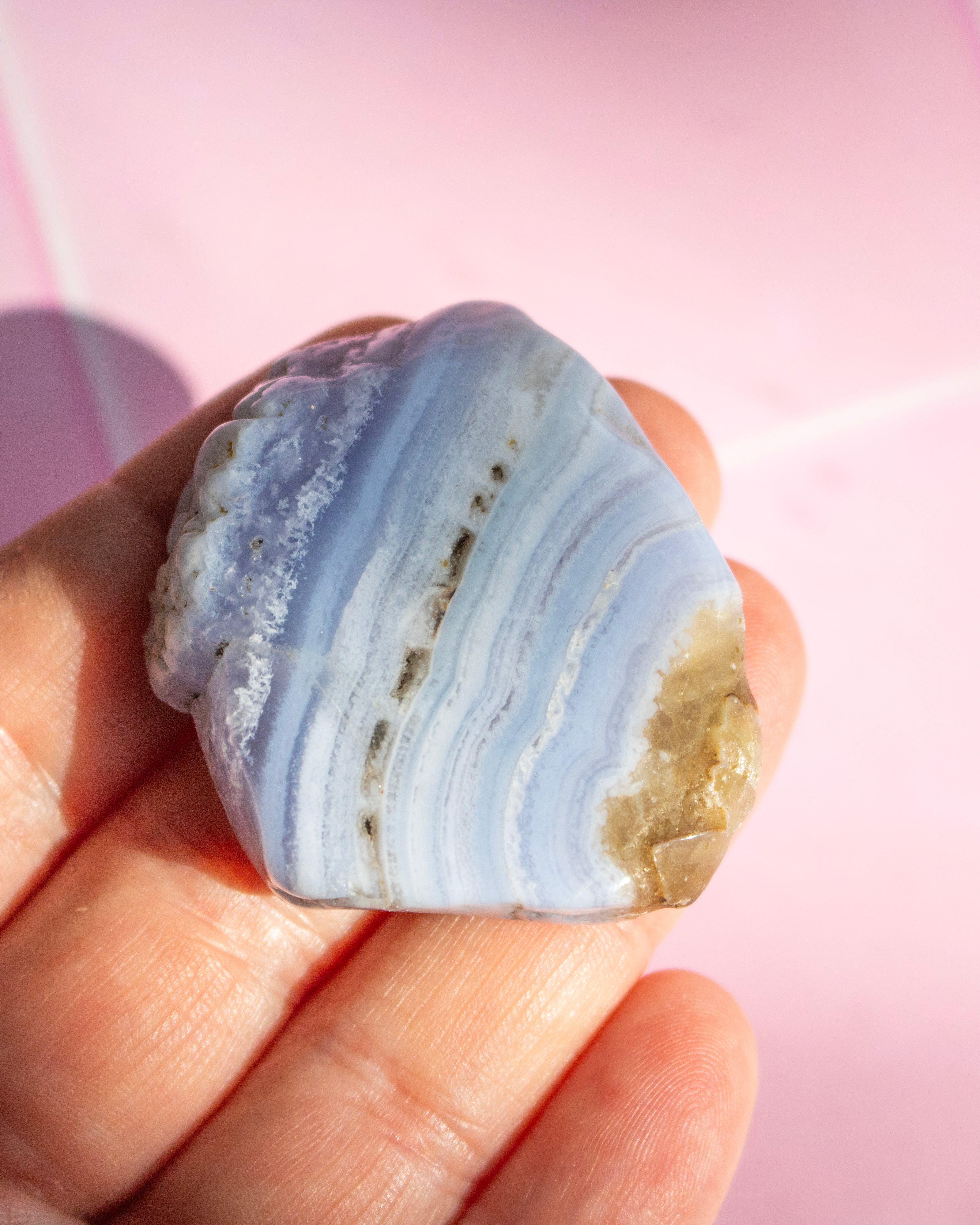 A hand holding a polished rectangular stone with natural wavy banding in pale lavender, light blue, and white, against a soft pink background.