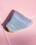 A polished slice of agate crystal with layered blue, purple, and white bands, resting on a soft pink background.