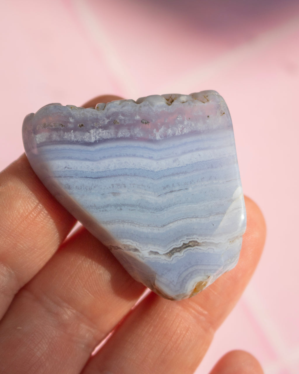 A polished piece of bandstone with horizontal light blue, lavender, and white stripes, held in a person's hand against a soft pink background.