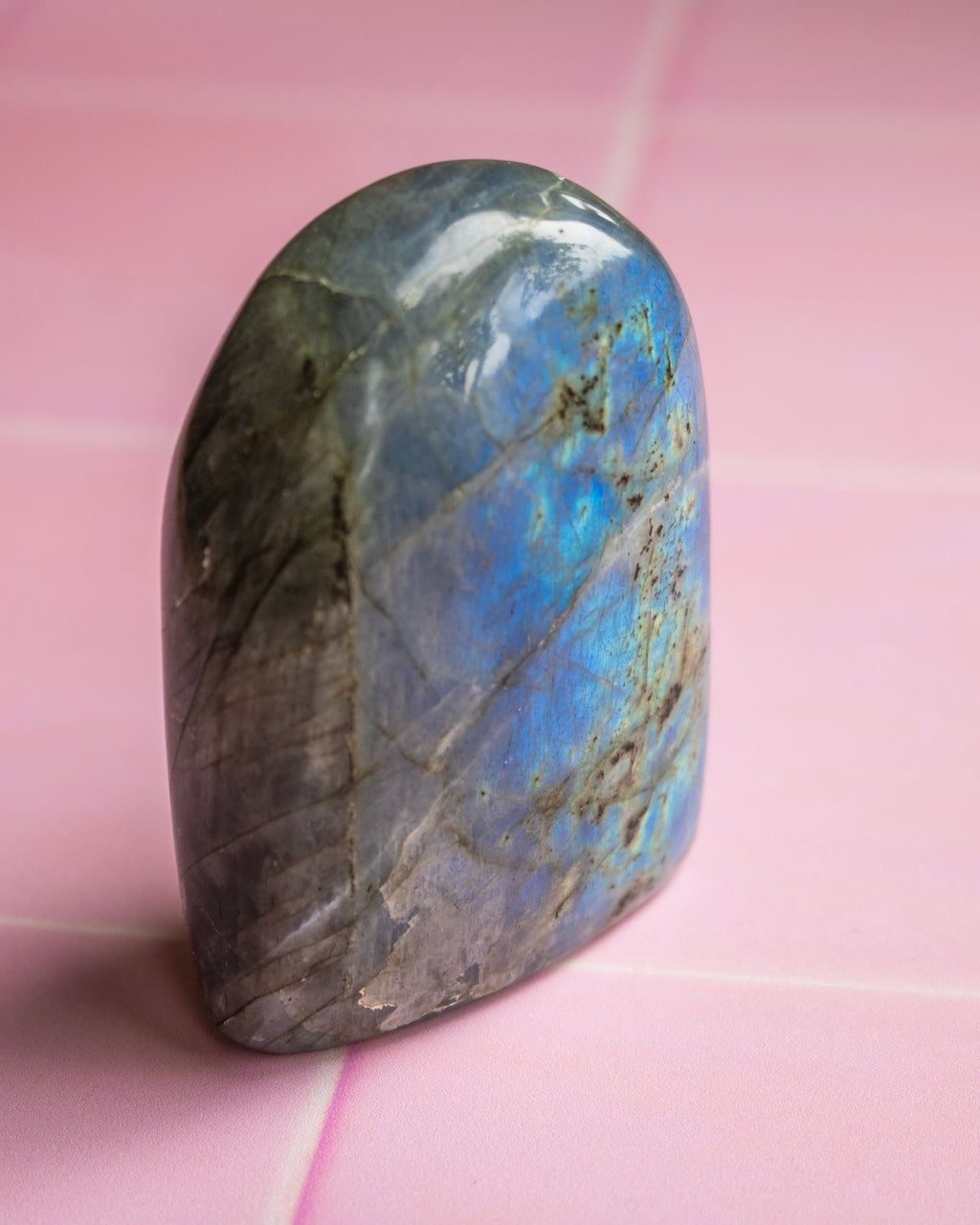 A polished labradorite crystal stone with a natural rounded shape, displaying a shimmering blue and gray play-of-color, resting on a pink tiled surface.