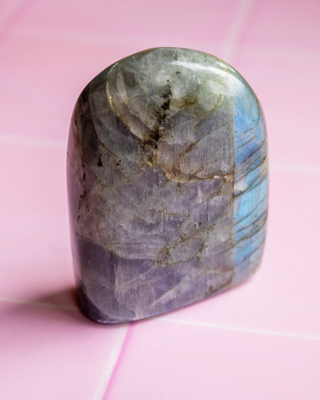 A polished labradorite crystal stone with natural gray, purple, and blue iridescent flashes, resting on a pink tiled surface.
