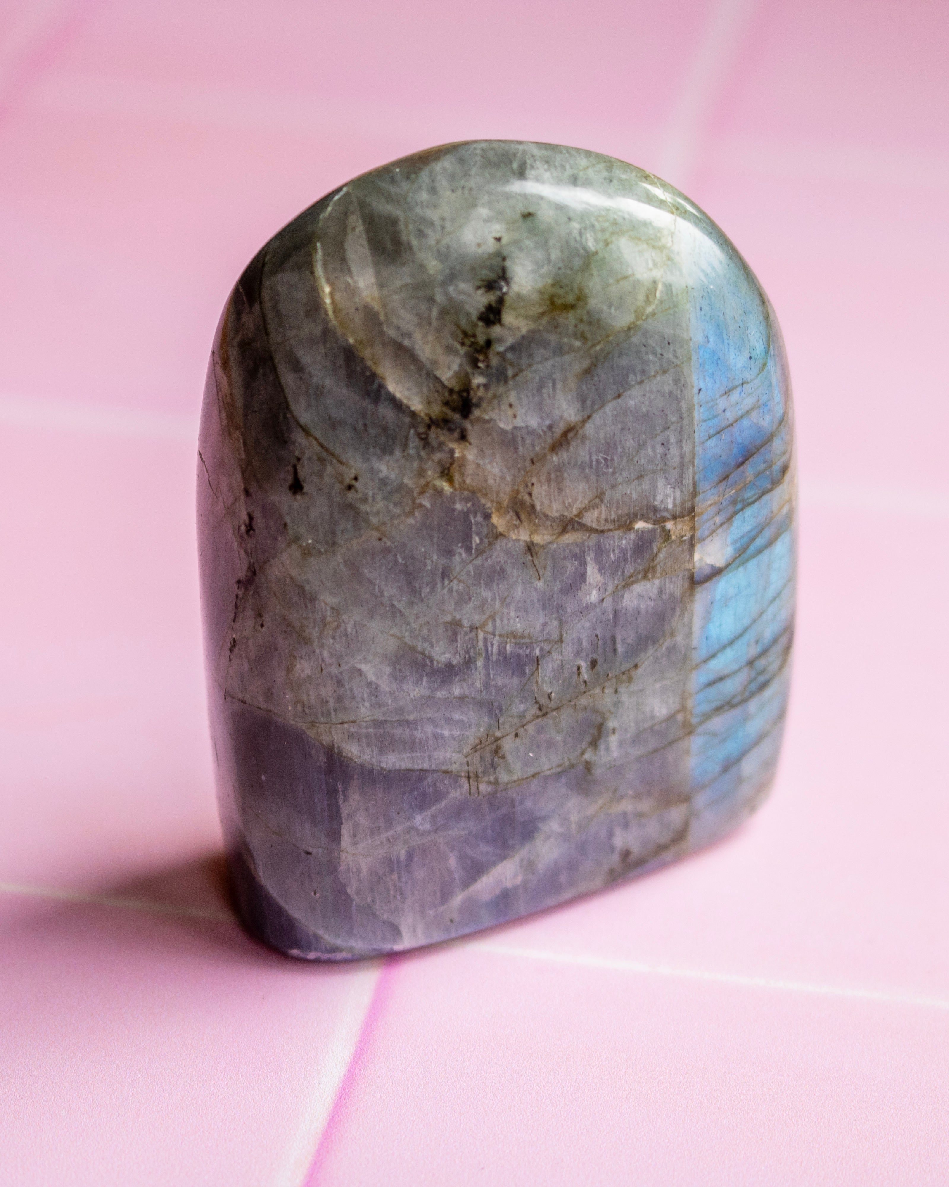 A polished labradorite crystal stone with natural gray, purple, and blue iridescent flashes, resting on a pink tiled surface.