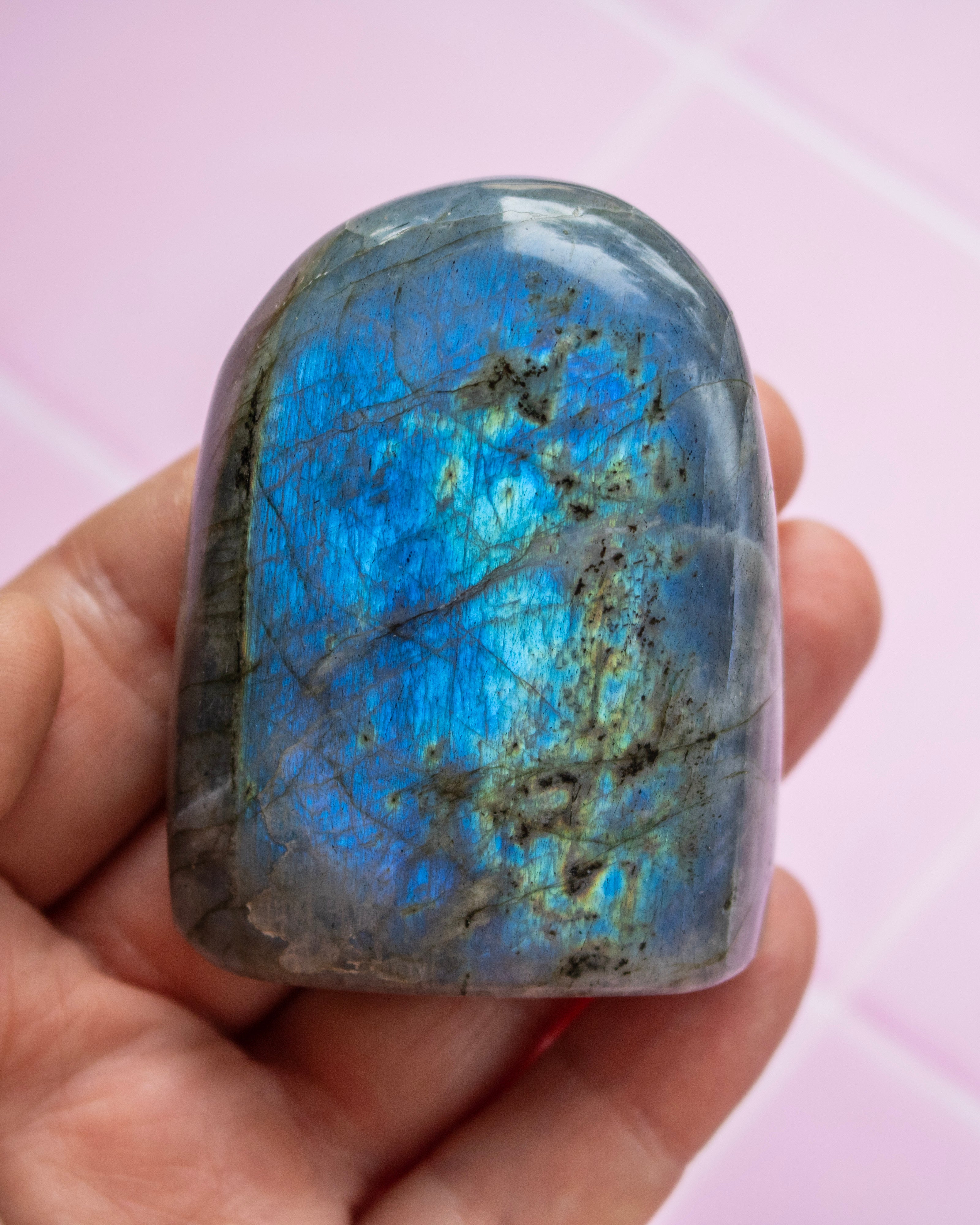 A polished labradorite stone held in a hand, showing a vibrant blue iridescent flash with gray and brown matrix patterns, against a soft pink tiled background.