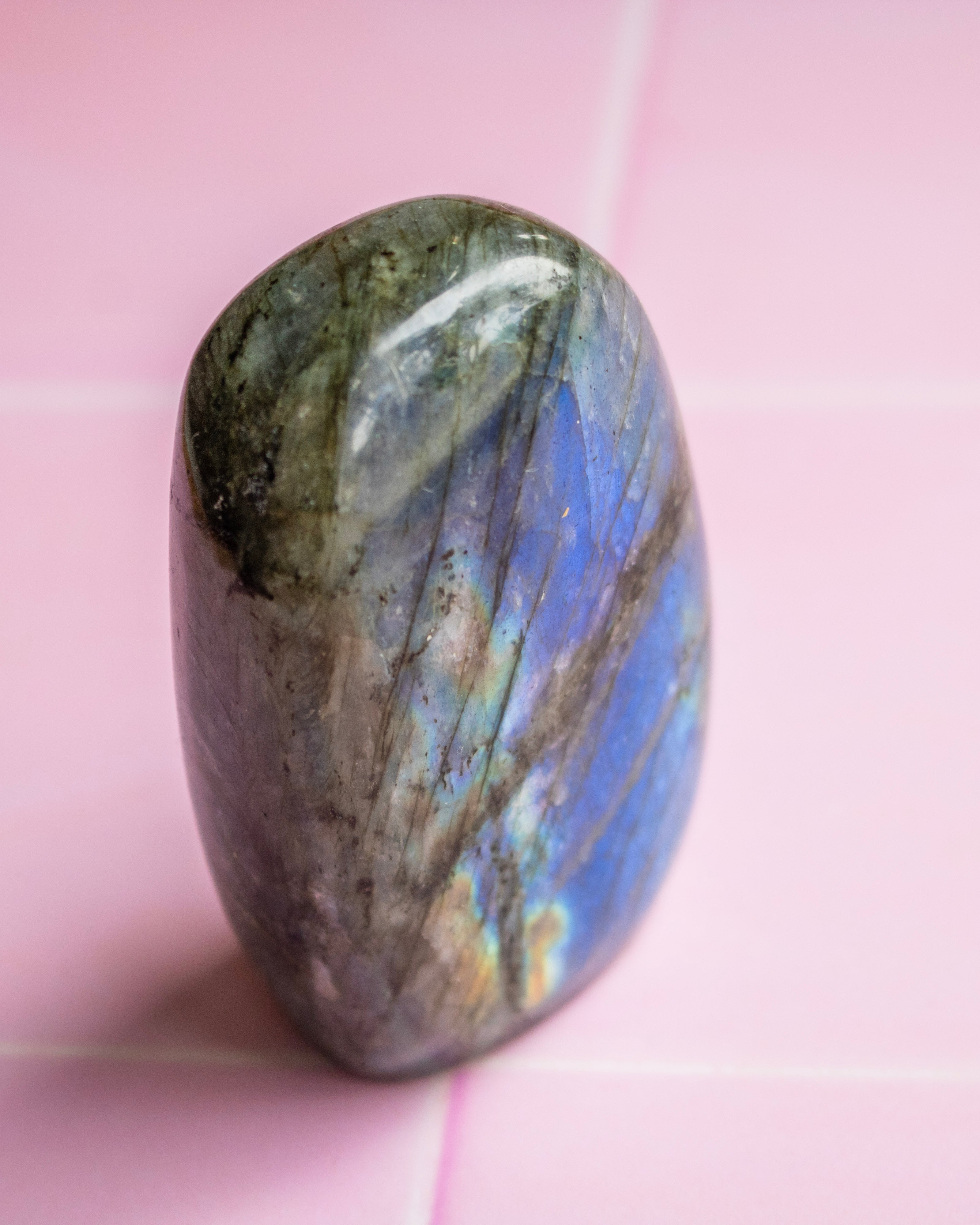 A polished labradorite tumbled stone with natural blue, gray, and green iridescence, standing upright on a light pink tiled surface.