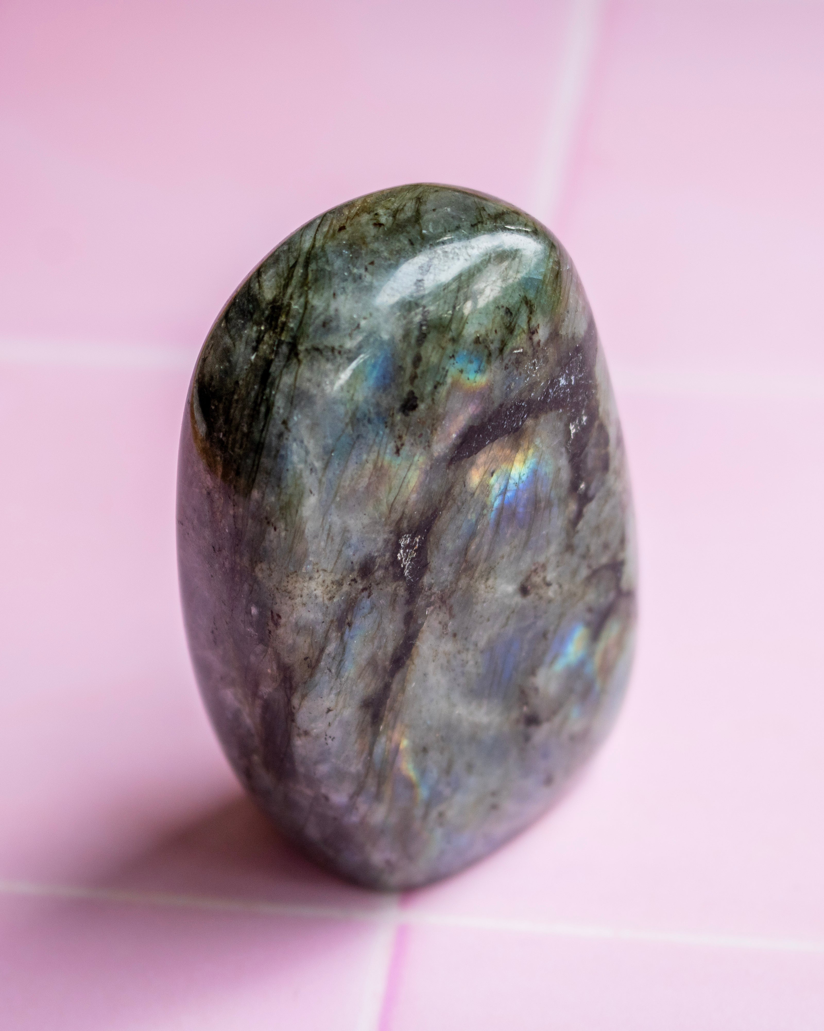 A polished tumbled labradorite crystal stone with iridescent blue and green flashes, standing upright on a light pink tiled surface.