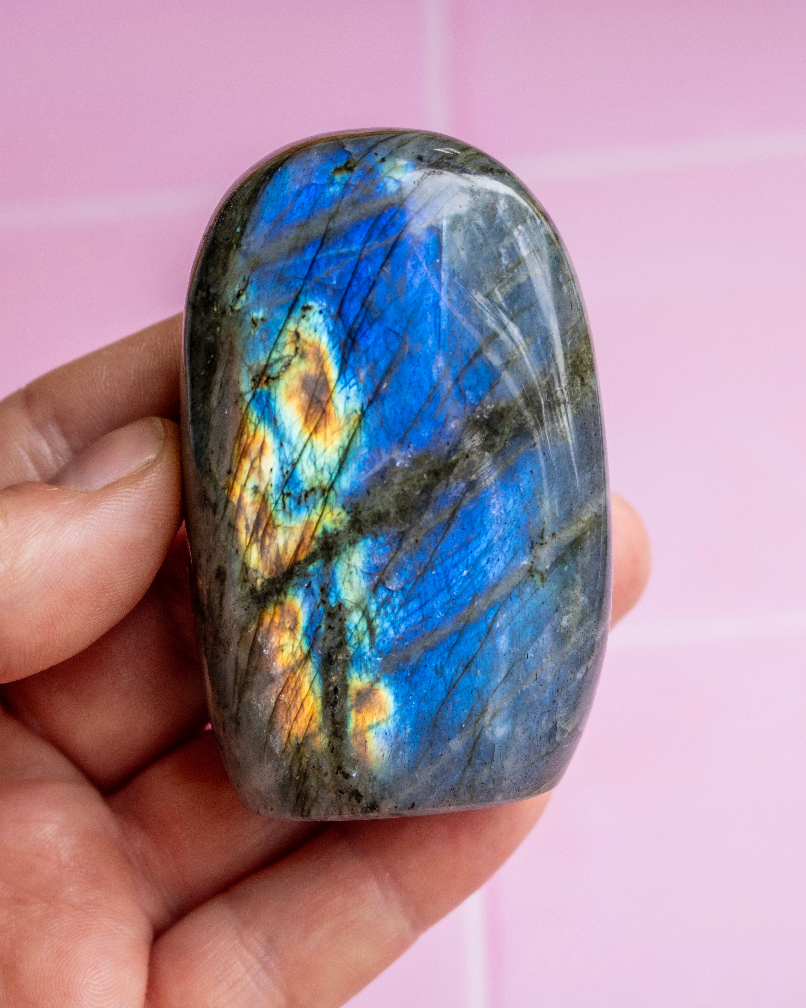 A hand holding a polished labradorite stone with vibrant blue, gold, and gray flashes, set against a soft pink background.
