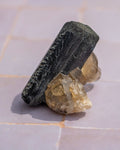 A natural mineral specimen featuring a large, dark gray to black rectangular crystal, likely a pyrite or similar silicate, resting on a cluster of translucent pale yellow and smoky quartz crystals, set against a light gray tiled background.