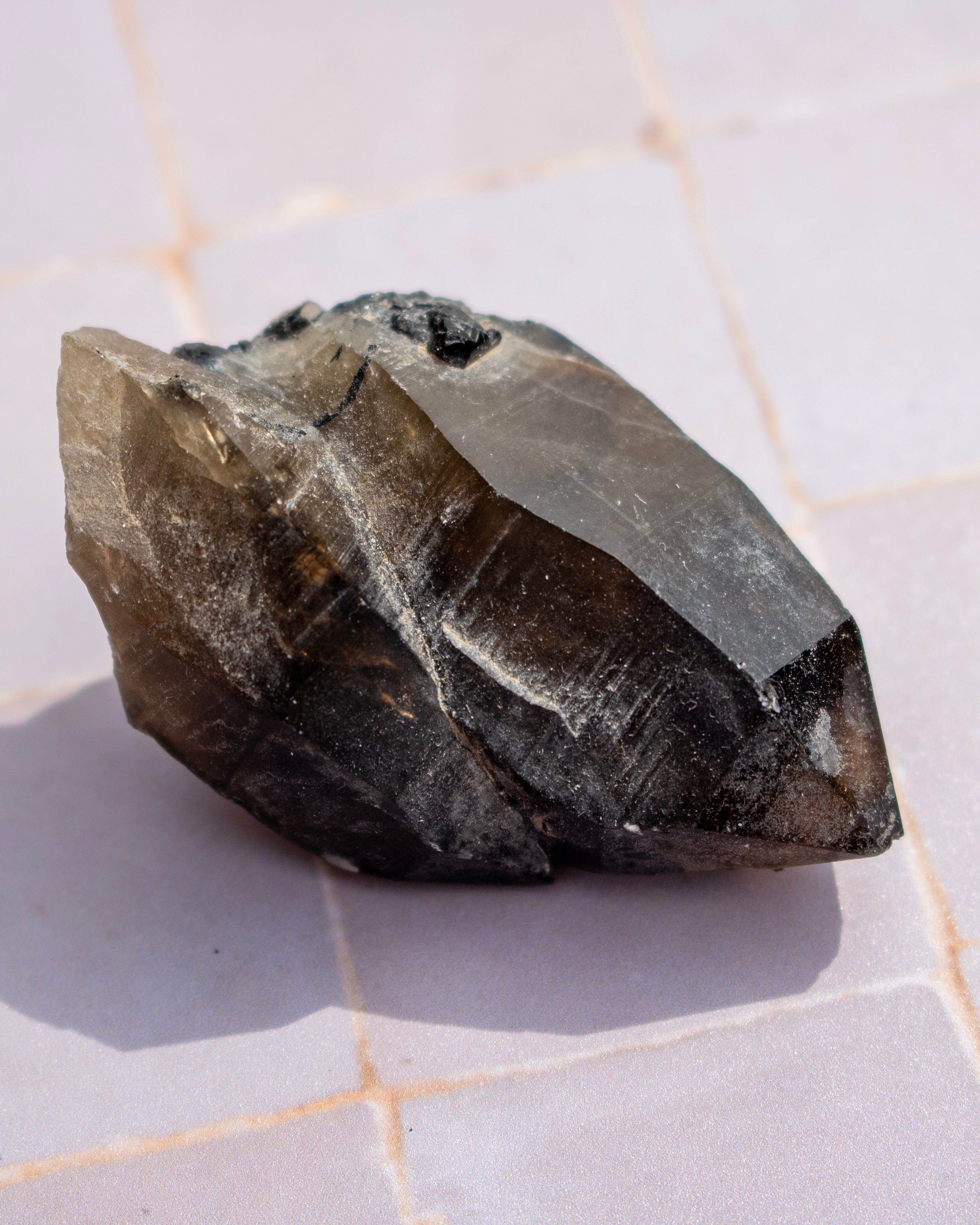 A rough, unpolished dark gray to black crystal or mineral specimen resting on a light gray tiled surface.
