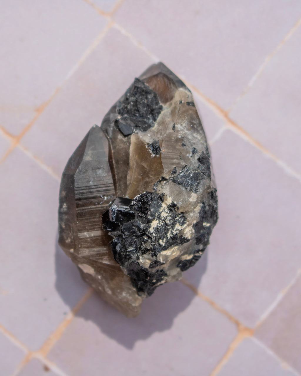 A raw mineral specimen showing a mix of smoky translucent quartz and dark metallic-looking crystals, resting on a light-colored tiled surface.