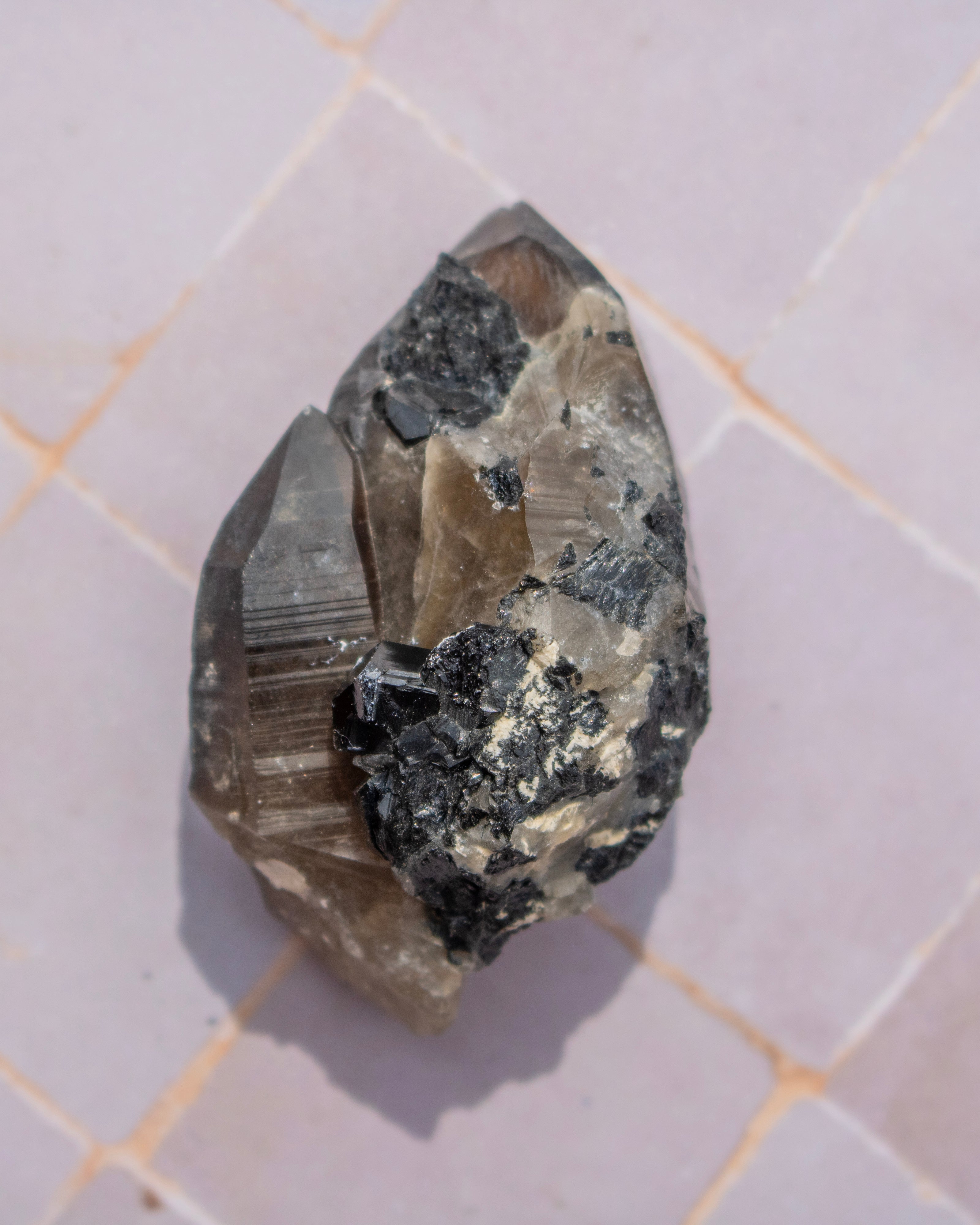 A raw mineral specimen showing a mix of smoky translucent quartz and dark metallic-looking crystals, resting on a light-colored tiled surface.