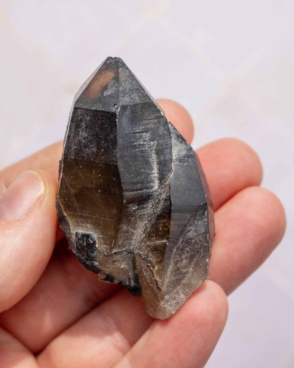 A hand holding a raw smoky quartz crystal point against a light, neutral background.