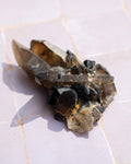 A natural mineral crystal cluster featuring prominent black points and smoky brown quartz crystal points, resting on a light-colored tiled surface.