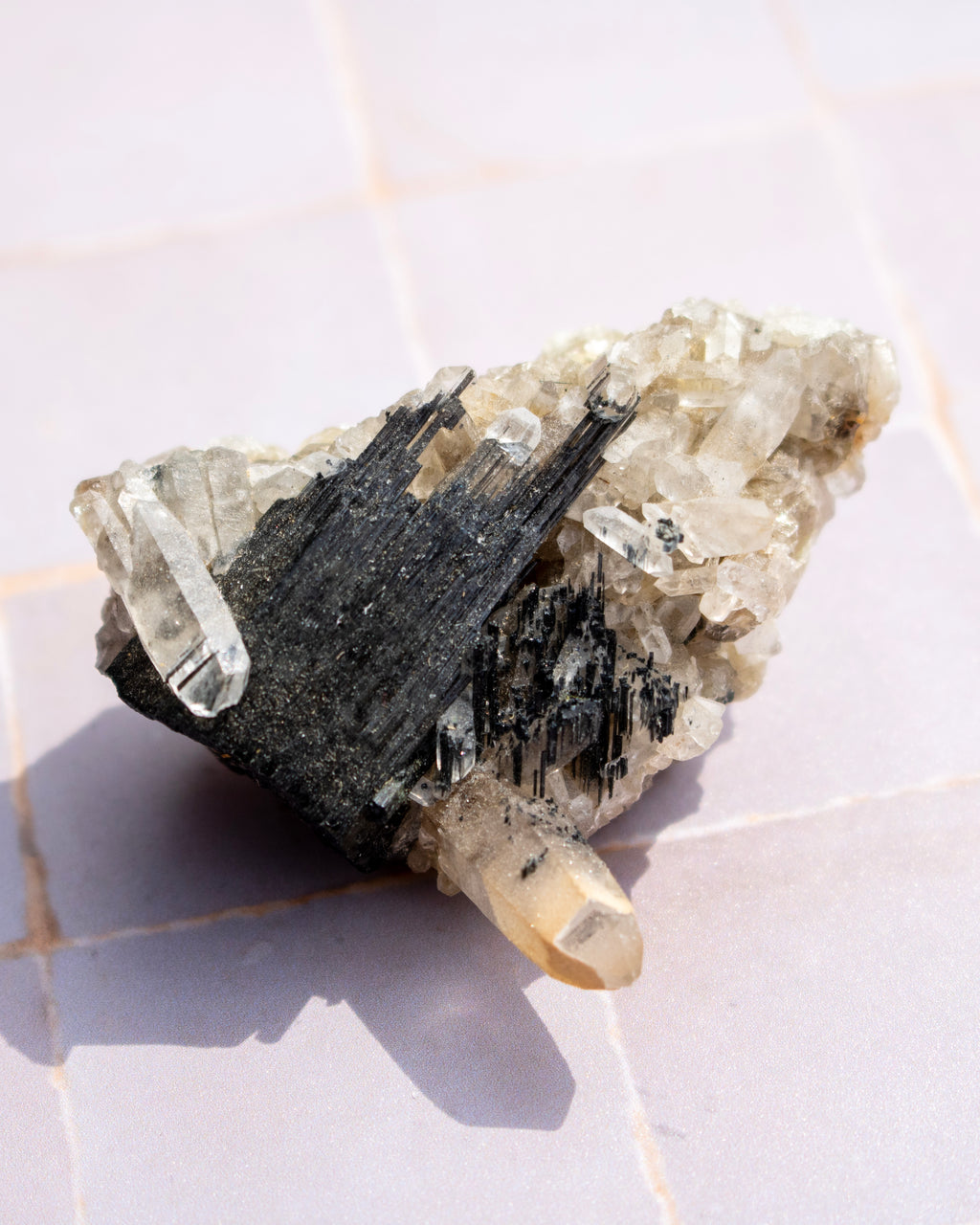 A natural mineral specimen featuring a cluster of clear quartz crystals and prominent black needle-like pyrite formations on a pale stone base, photographed on a light tiled surface.