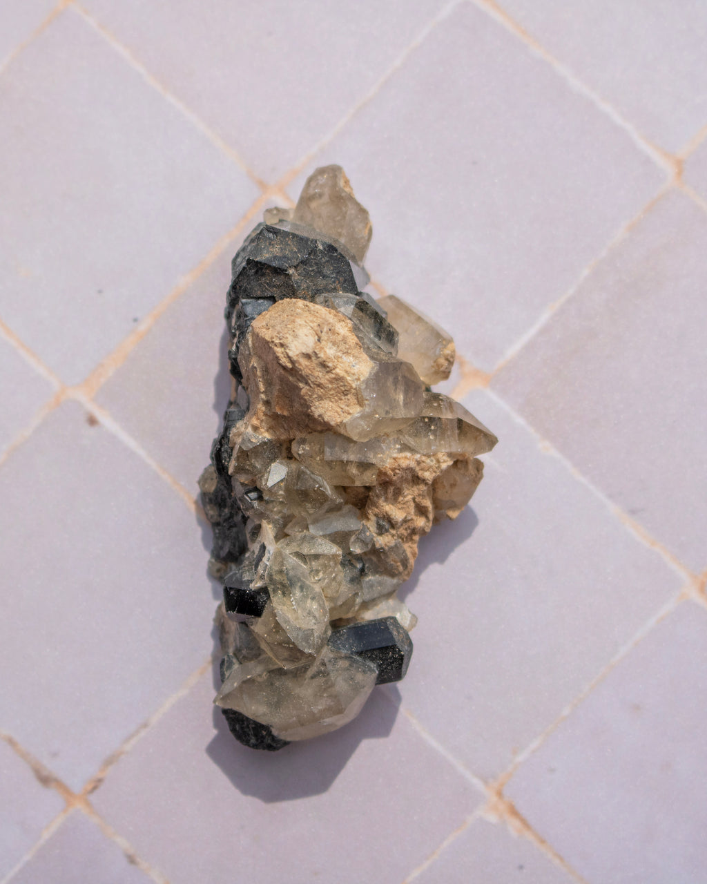 A raw mineral specimen with mixed gray, beige, and black crystalline textures, photographed on a light gray tiled surface.