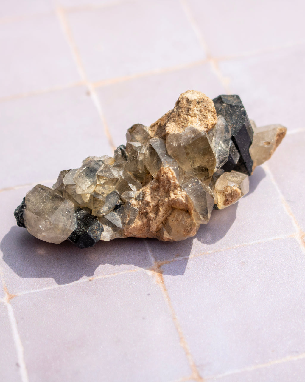 A natural crystal cluster with faceted clear and smoky quartz-like stones, tan mineral deposits, and dark gray to black sections, photographed on a light tiled surface.
