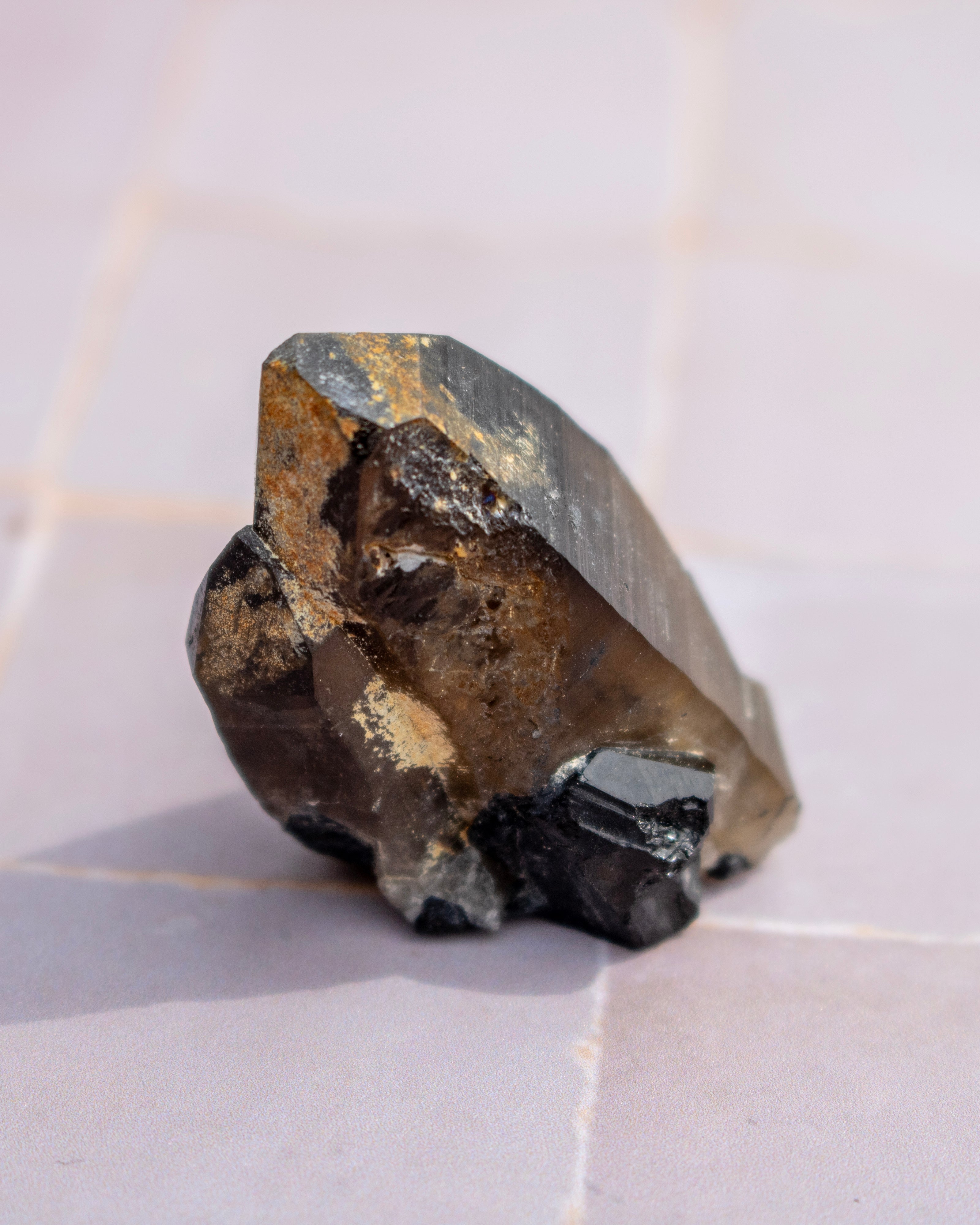 A raw mineral specimen with a faceted dark gray crystal structure, showing prominent brown and gold crystalline inclusions, resting on a light beige tiled surface.
