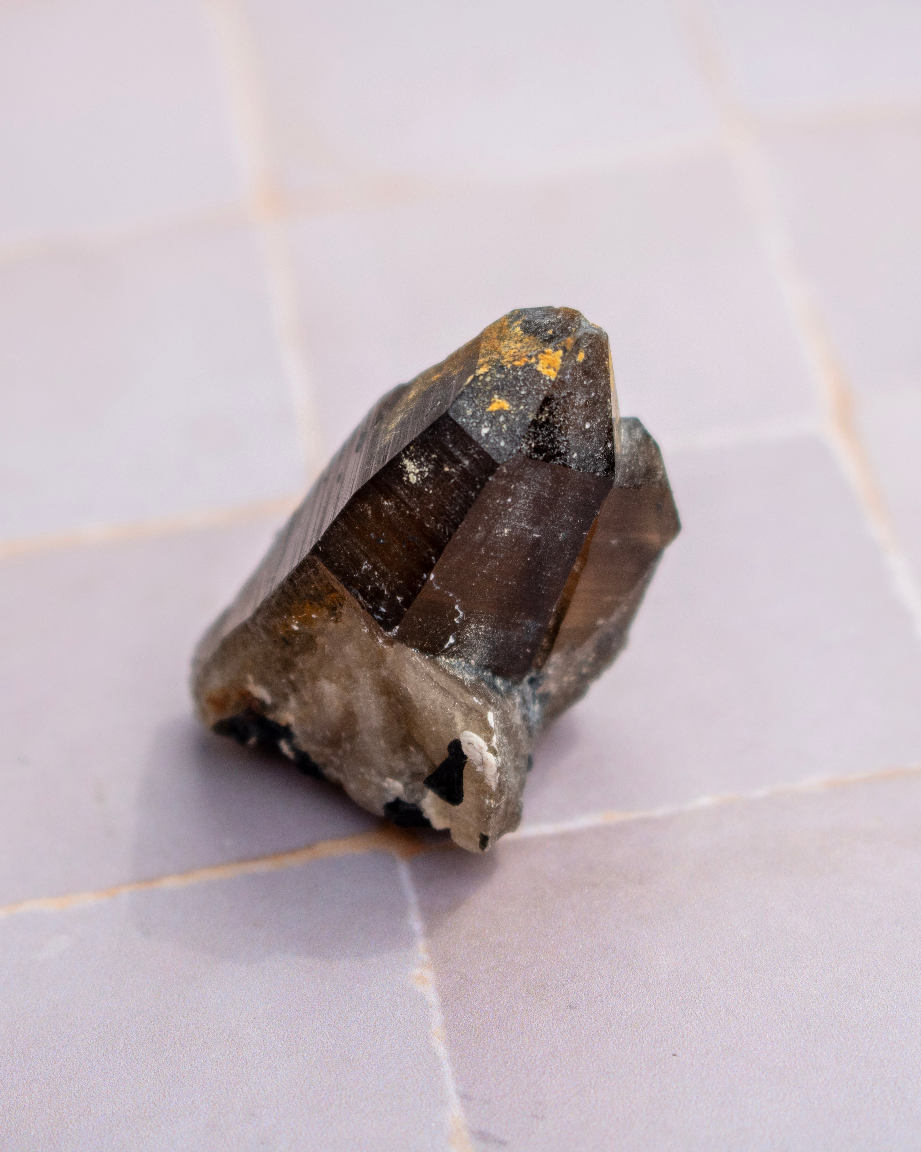A raw smoky quartz crystal cluster with dark brown and black translucent sections and small gold inclusions, resting on a light gray tiled surface.