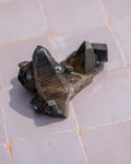 A cluster of natural smoky quartz crystal points with dark gray to black striated matrix, resting on a light pinkish-beige tiled surface.