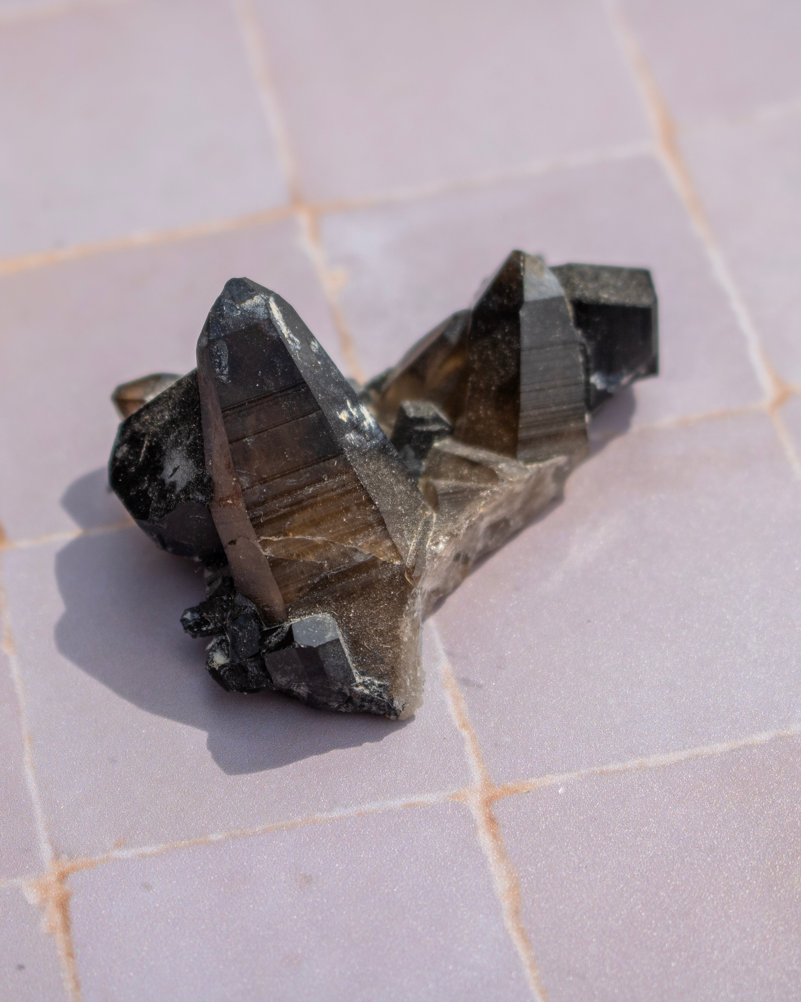 A cluster of natural smoky quartz crystal points with dark gray to black striated matrix, resting on a light pinkish-beige tiled surface.