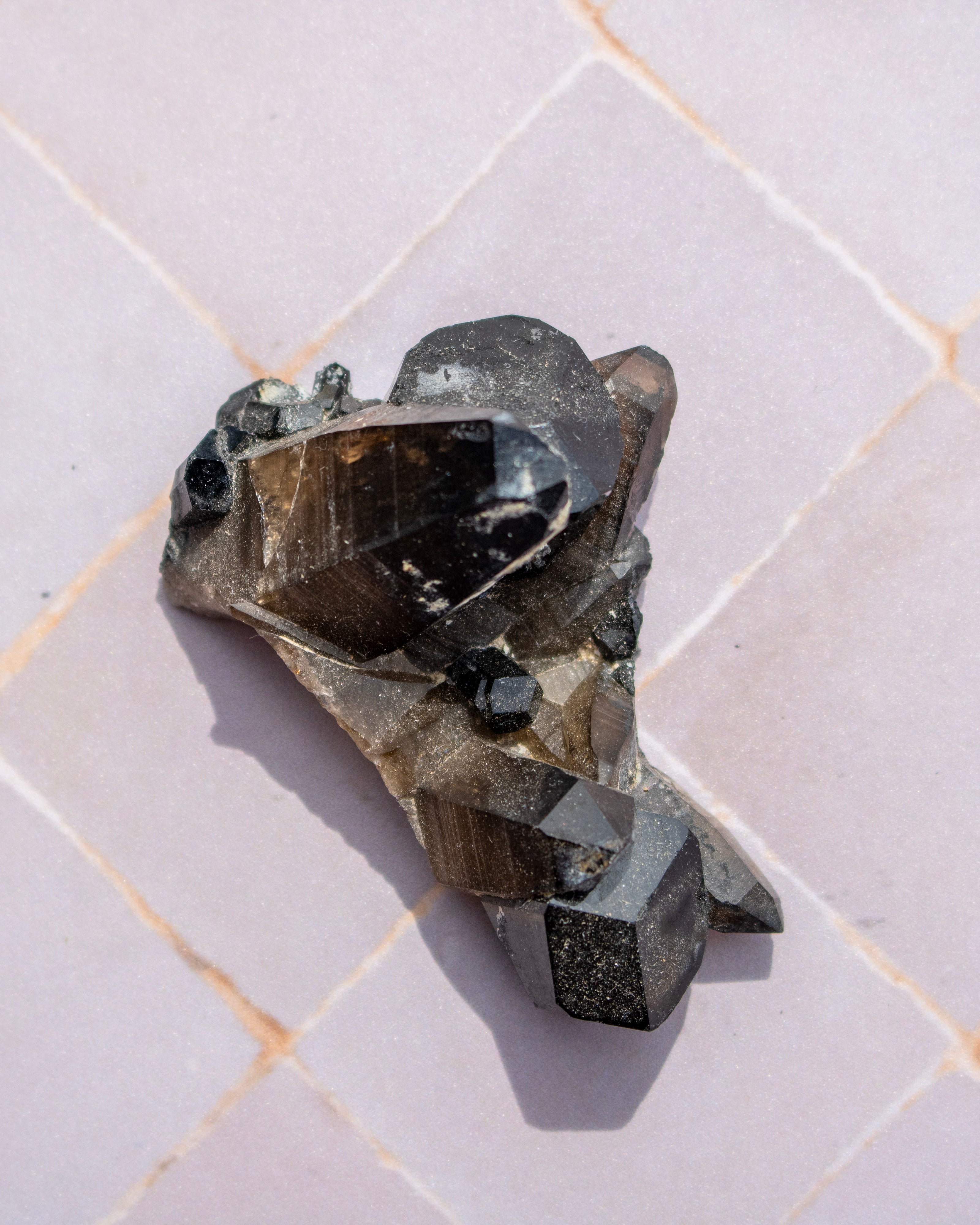 A natural smoky quartz crystal cluster with dark gray and black facets, resting on a light beige tiled surface.