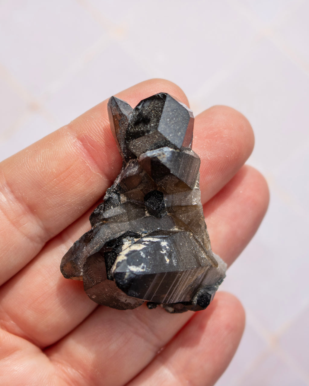 A hand holding a raw smoky quartz crystal cluster with dark gray to black facets against a light tiled background.