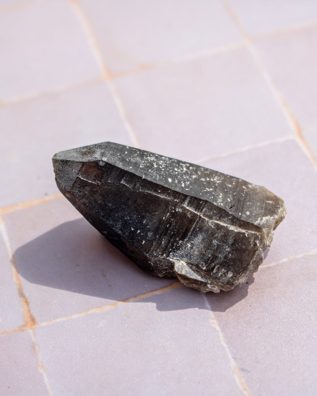 A raw dark gray mineral crystal with natural rough edges and a pointed tip, resting on a light gray tiled surface.