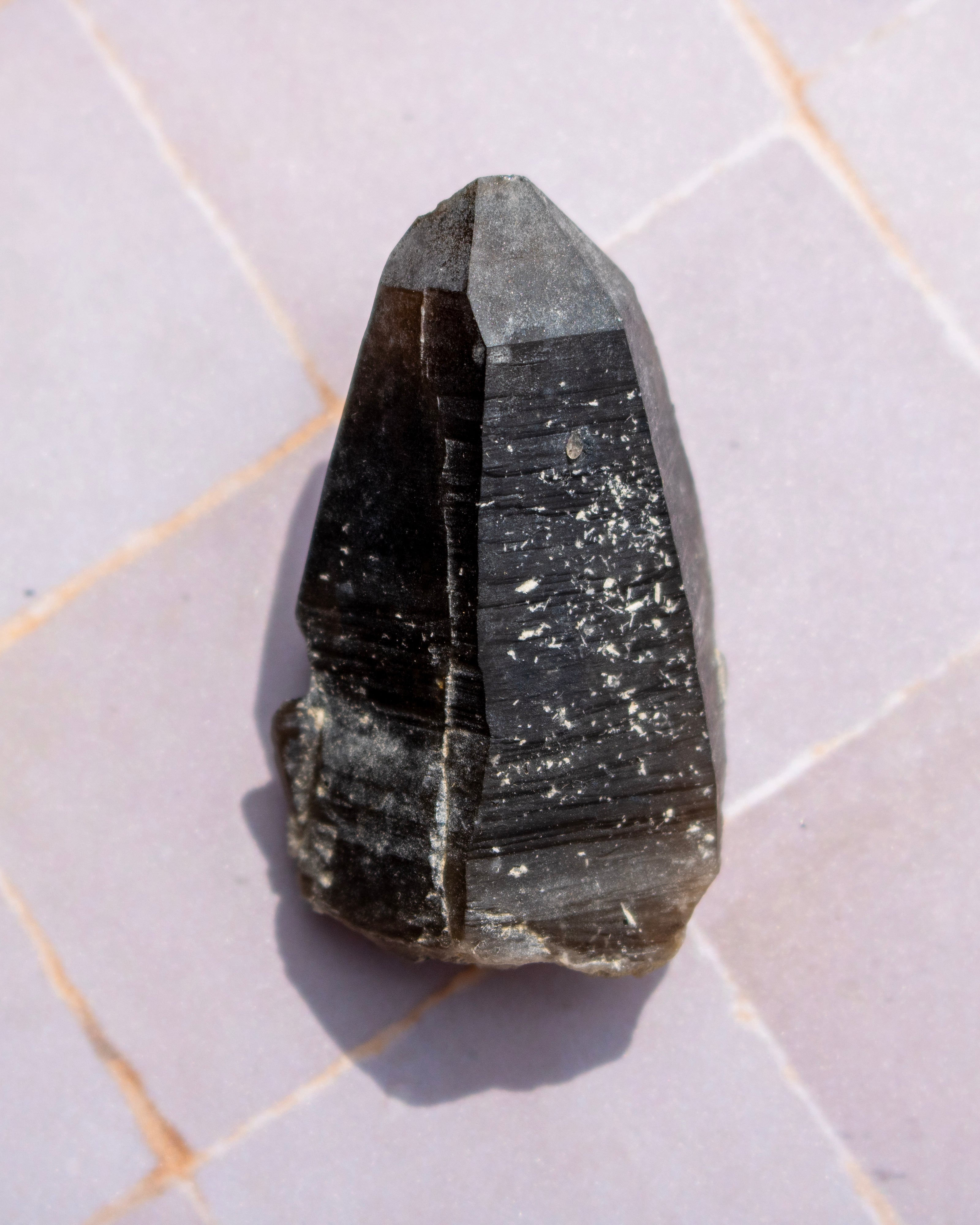 A natural dark gray to black smoky quartz crystal point resting on a light gray tiled surface.