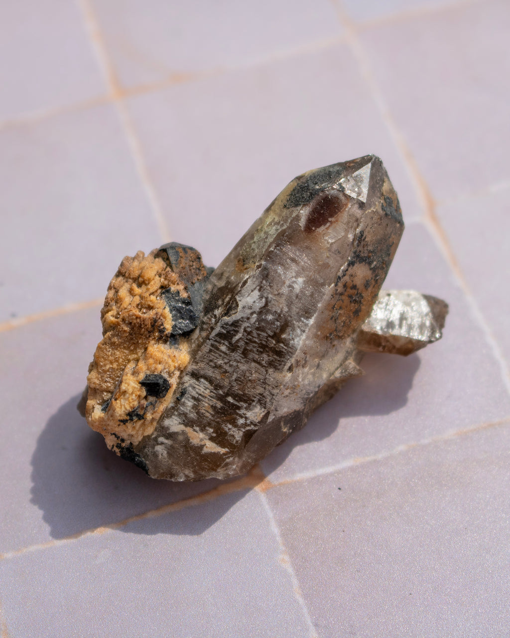 A raw smoky quartz crystal cluster with a rough, earthy matrix and a small metallic silver crystal point, resting on a light gray tiled surface.