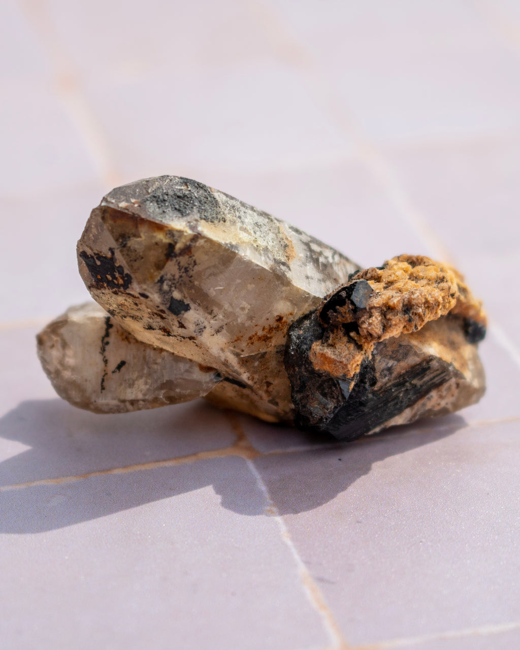 A cluster of natural crystals and mineral specimens, including a prominent clear quartz crystal with dark matrix and a rough brown textured piece, resting on light-colored square tiles.