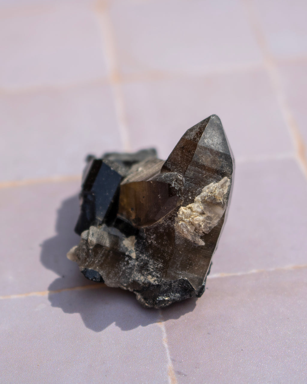 A natural smoky quartz crystal cluster with dark brown and black tones, resting on a light pinkish-beige tiled surface.