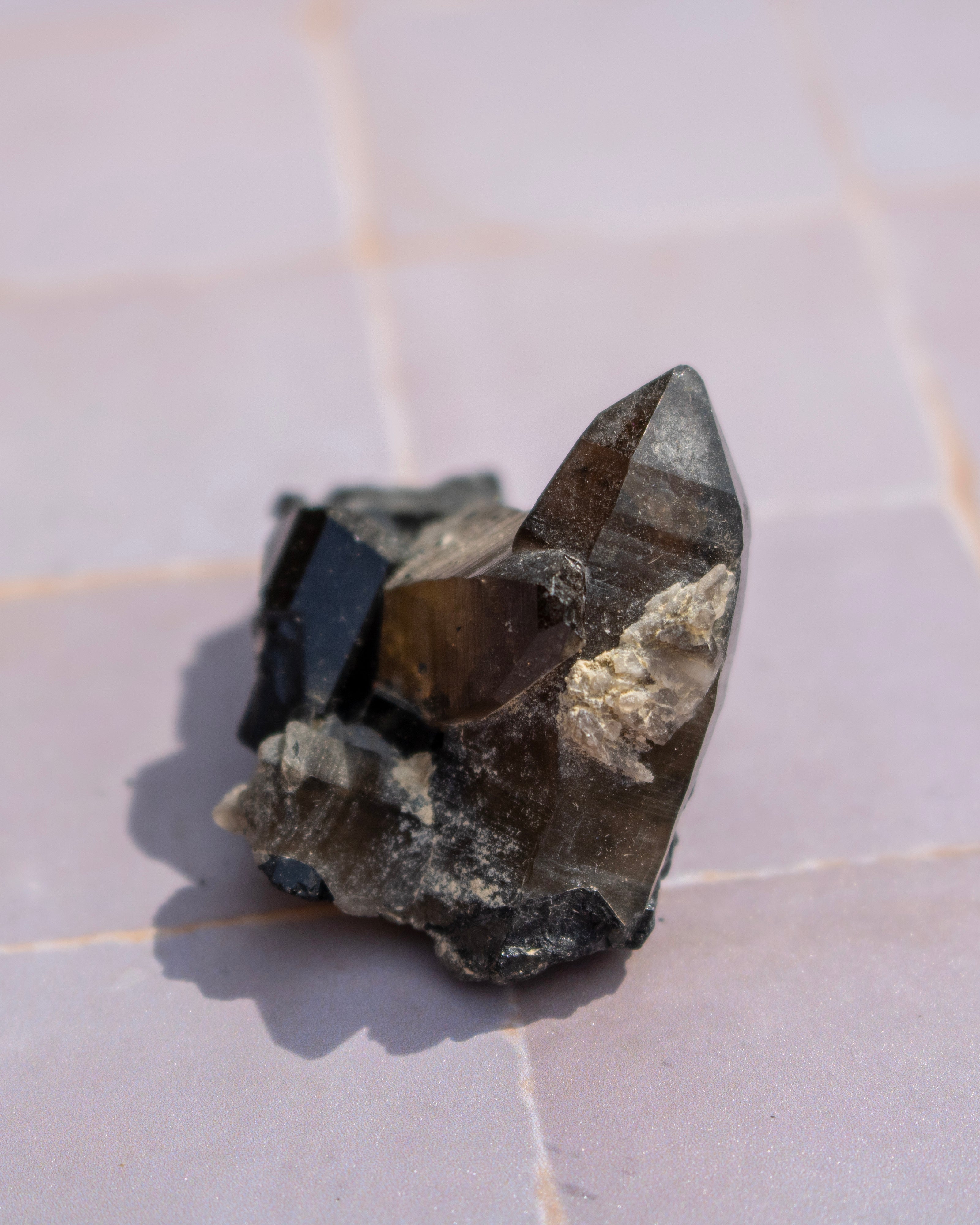 A natural smoky quartz crystal cluster with dark brown and black tones, resting on a light pinkish-beige tiled surface.