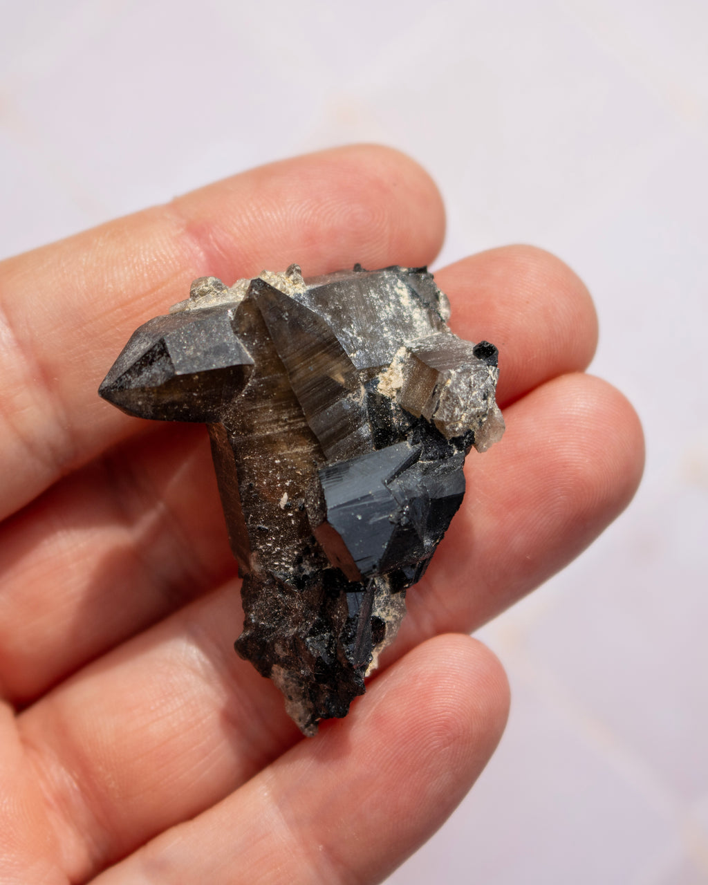 A hand holding a raw mineral or crystal specimen with dark gray to black faceted crystals and some lighter gray matrix, photographed against a pale background.