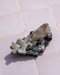 A natural crystal cluster featuring a prominent translucent smoky quartz point on a dark, rough mineral matrix, resting on a light-colored tiled surface.