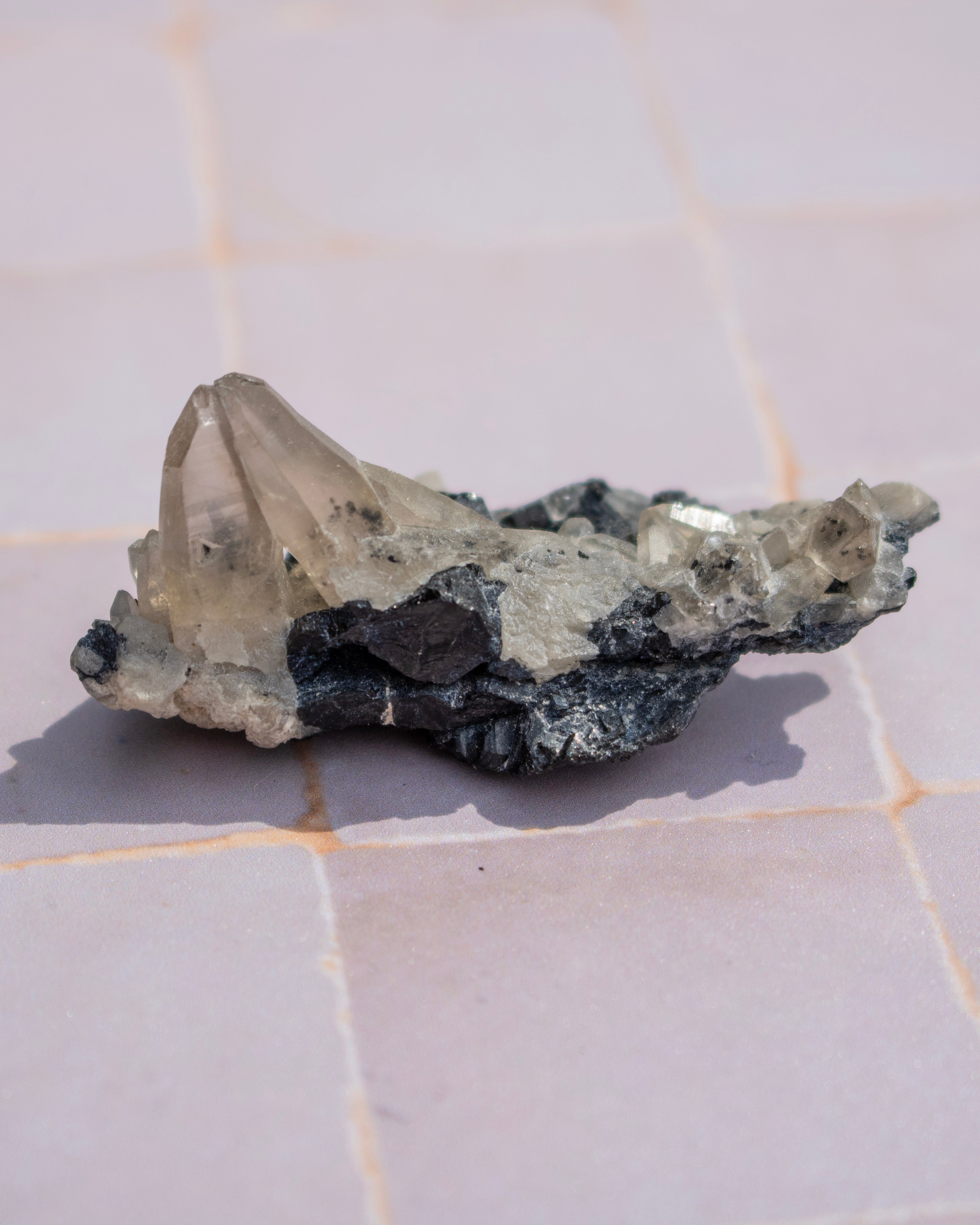 A raw mineral specimen featuring a prominent translucent clear quartz crystal cluster on a dark, textured black and gray matrix, resting on a light pink tiled surface.