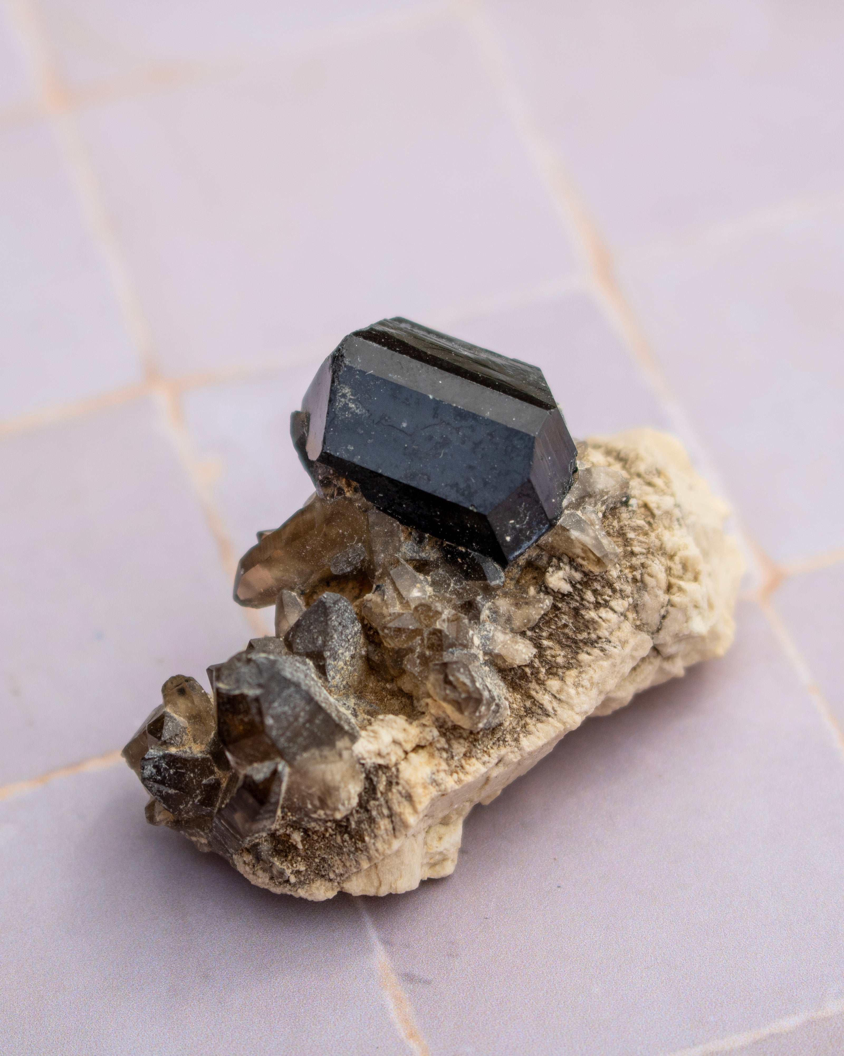 A faceted black gemstone, likely a cuboid or octagonal cut, displayed on top of a textured beige and brown mineral matrix, set against a light pink tiled background.