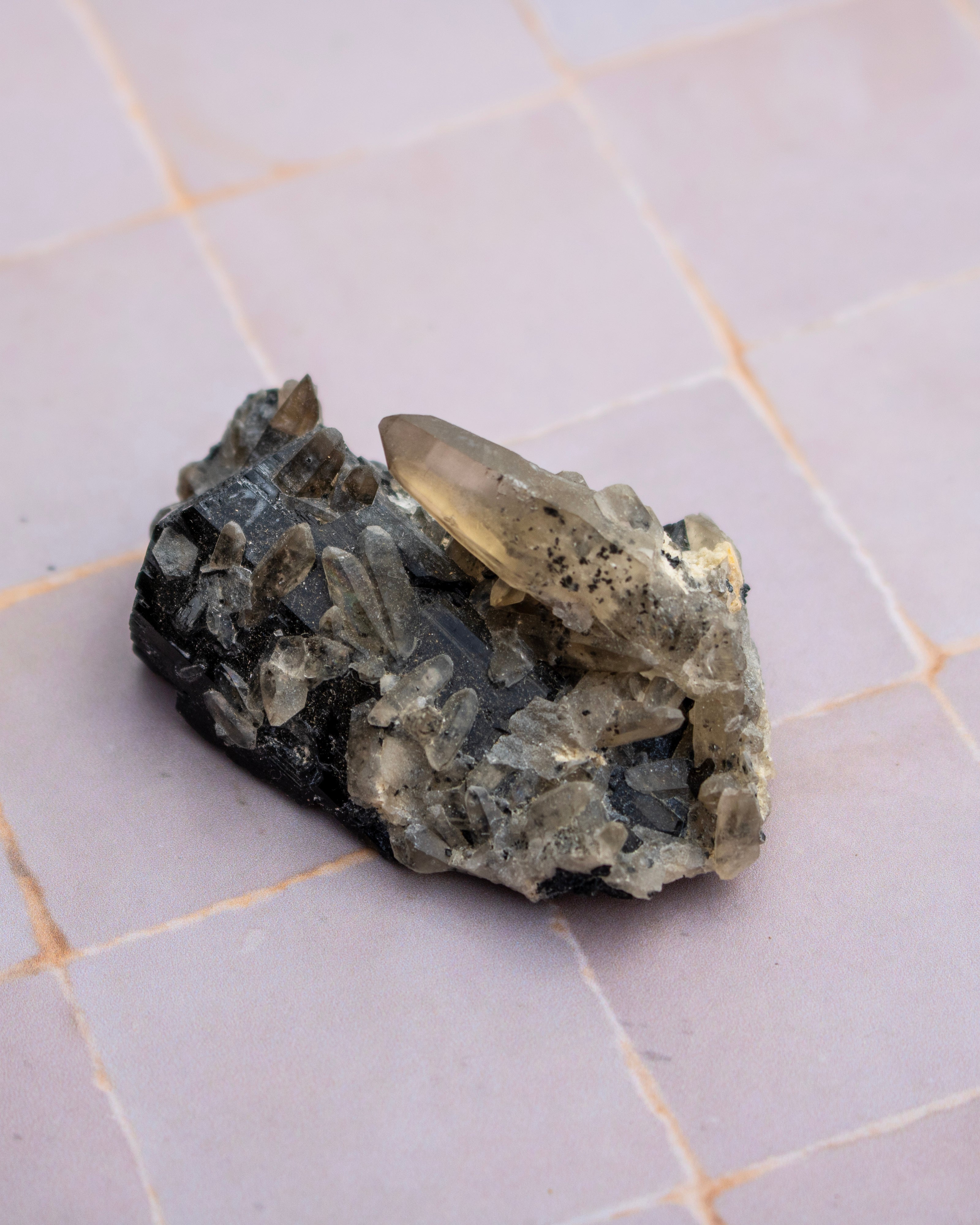 A raw smoky quartz crystal cluster with a rough, earthy matrix and a small metallic silver crystal point, resting on a light gray tiled surface.