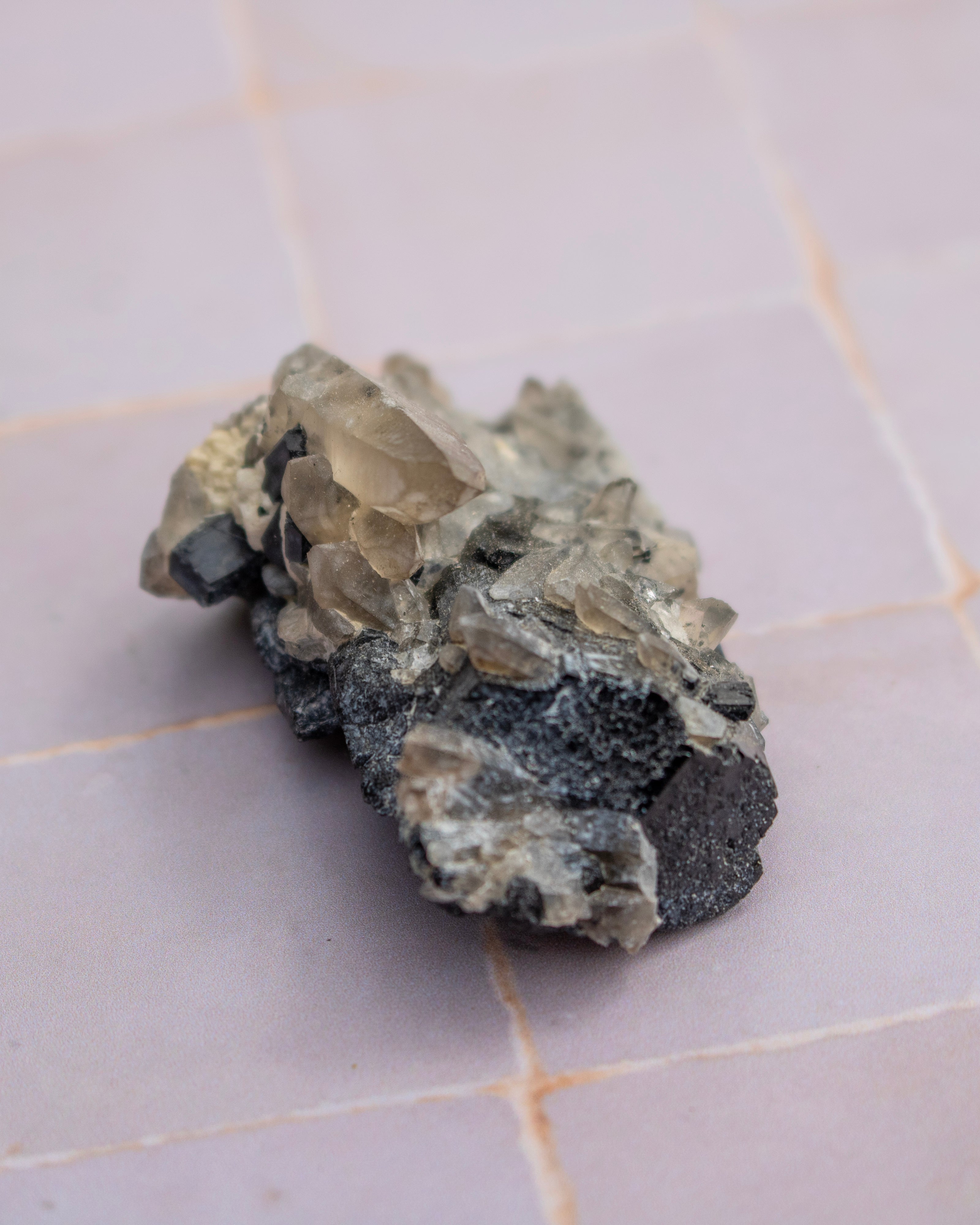 A raw smoky quartz crystal cluster with a rough, earthy matrix and a small metallic silver crystal point, resting on a light gray tiled surface.