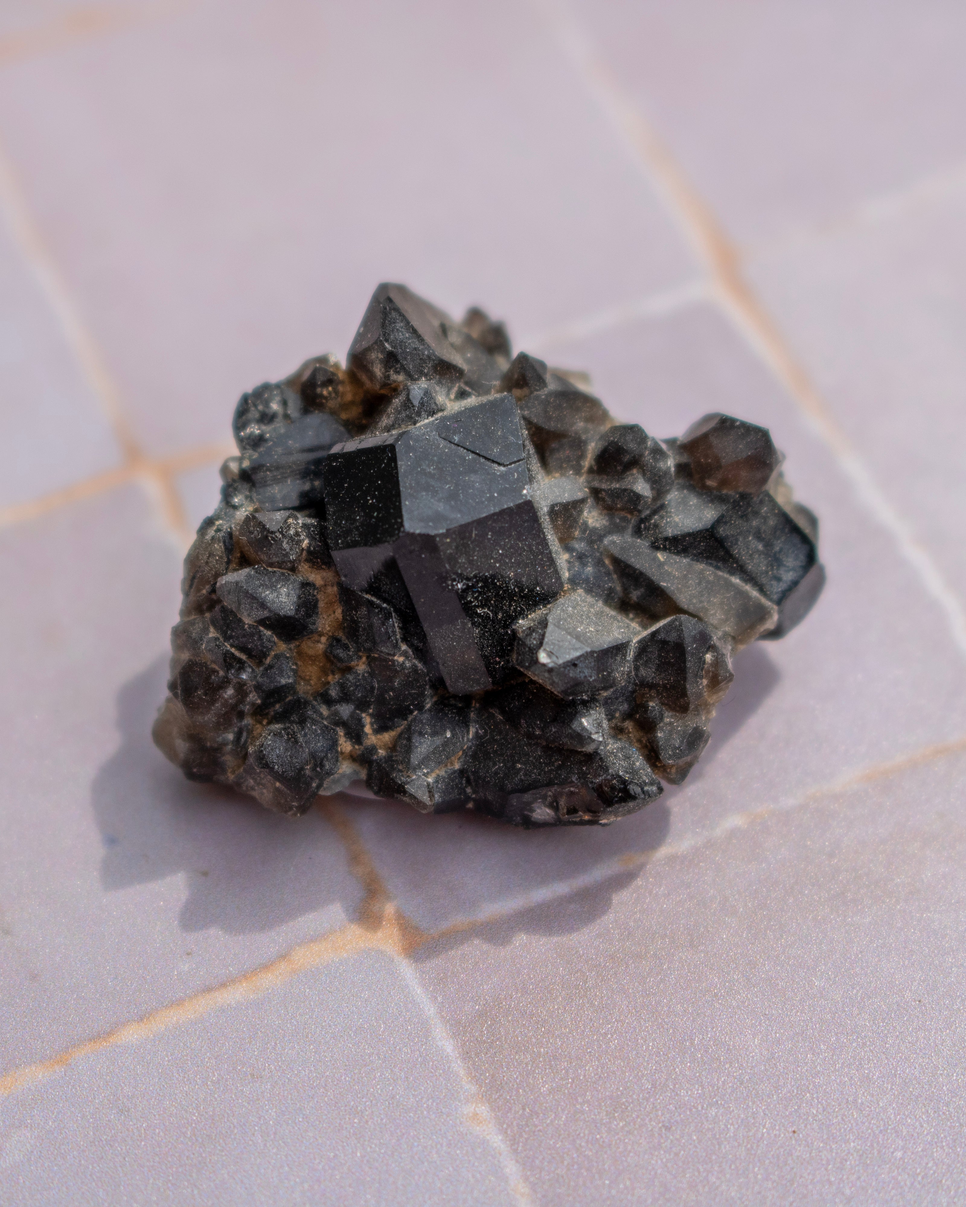 A raw crystal cluster of metallic dark gray to black cubic gemstones, likely pyrite, resting on a light gray tiled surface.