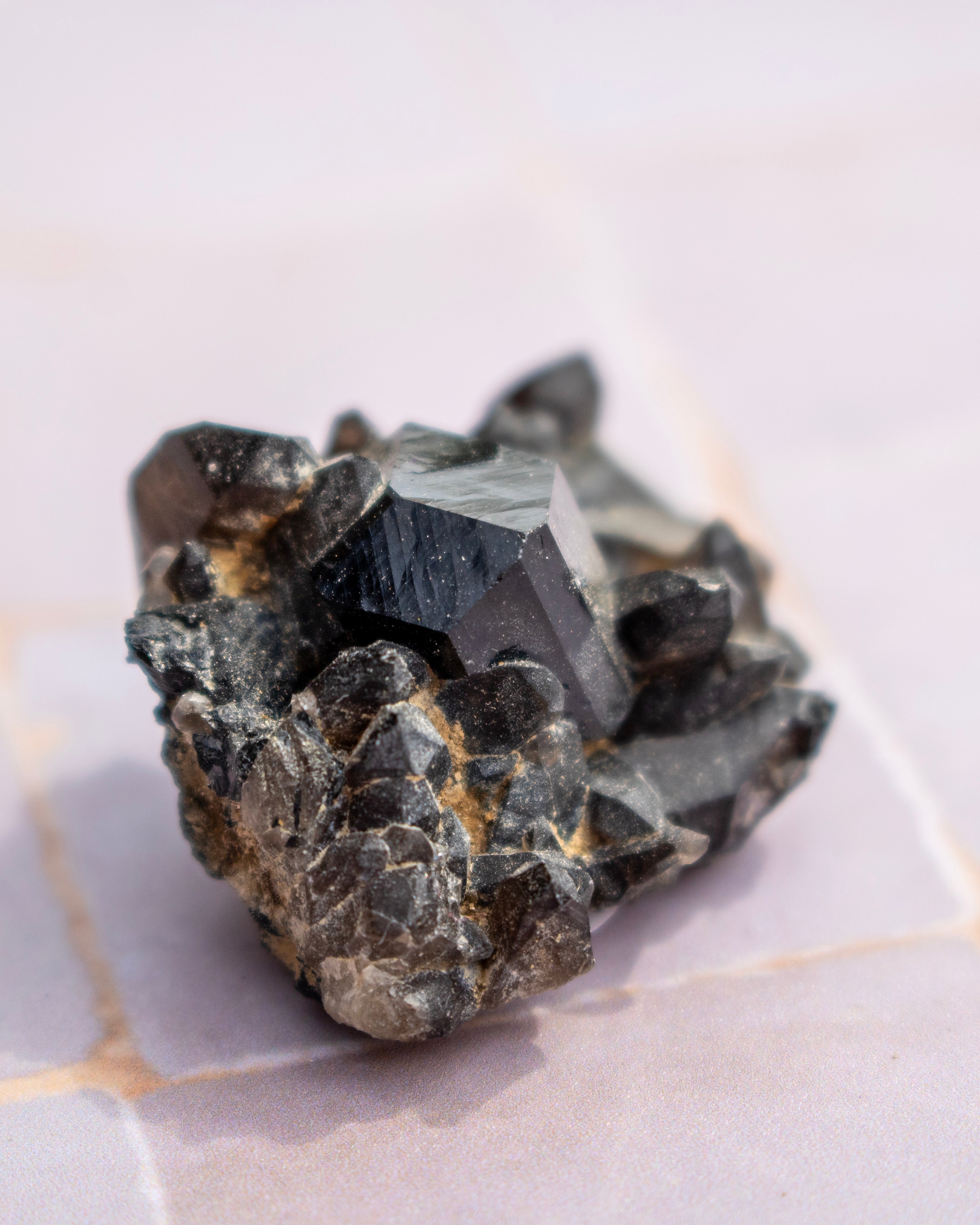 A rough black mineral crystal cluster with a prominent dark gray cuboid crystal, resting on a light pink-tinted tiled surface.