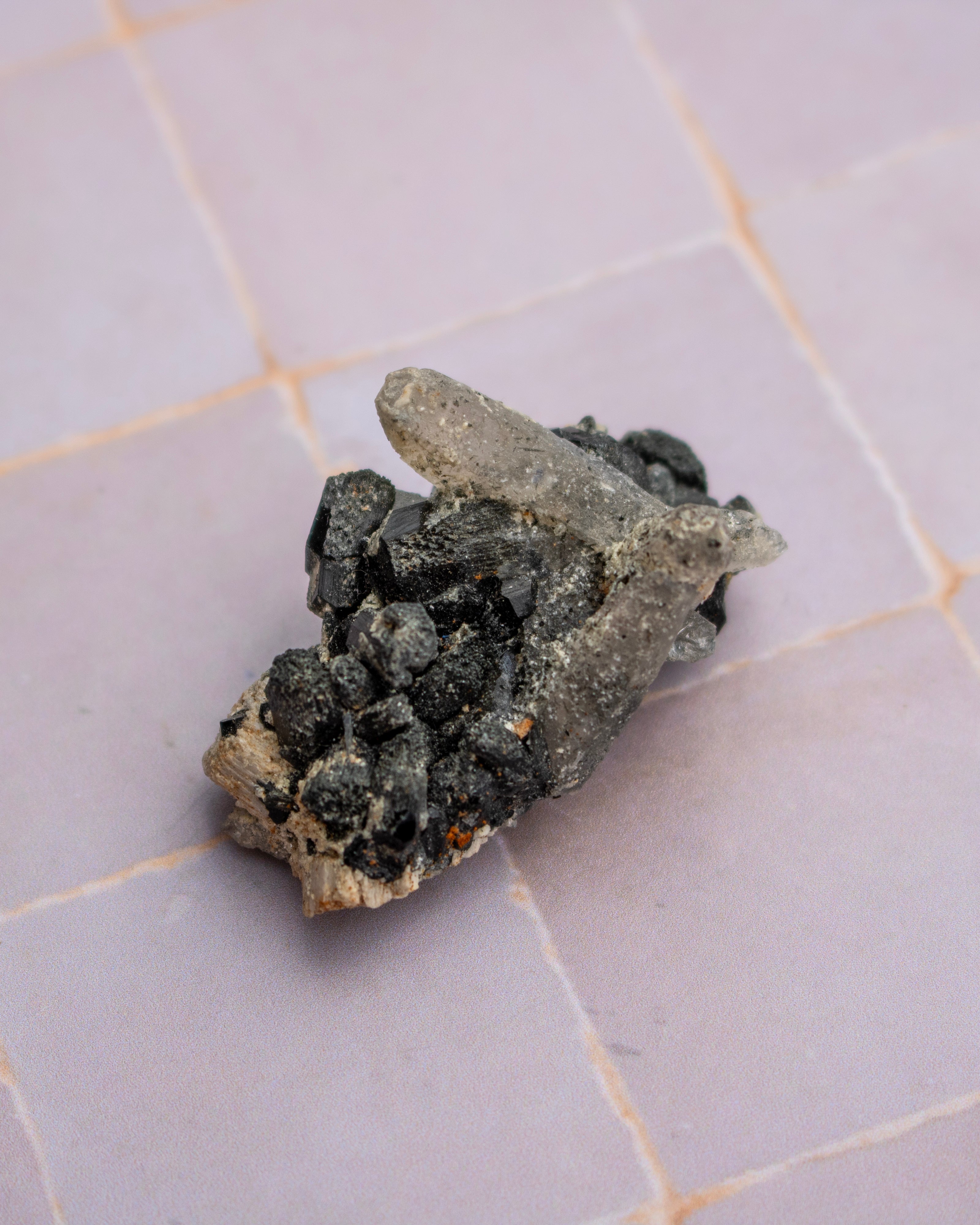 A natural mineral specimen featuring a cluster of dark, porous black crystals on a light gray and beige matrix, resting on a light purple tiled surface.
