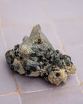A natural mineral specimen featuring a pointed crystal cluster with black and gray crystals set in a sandy beige matrix, resting on a light-colored tiled surface.