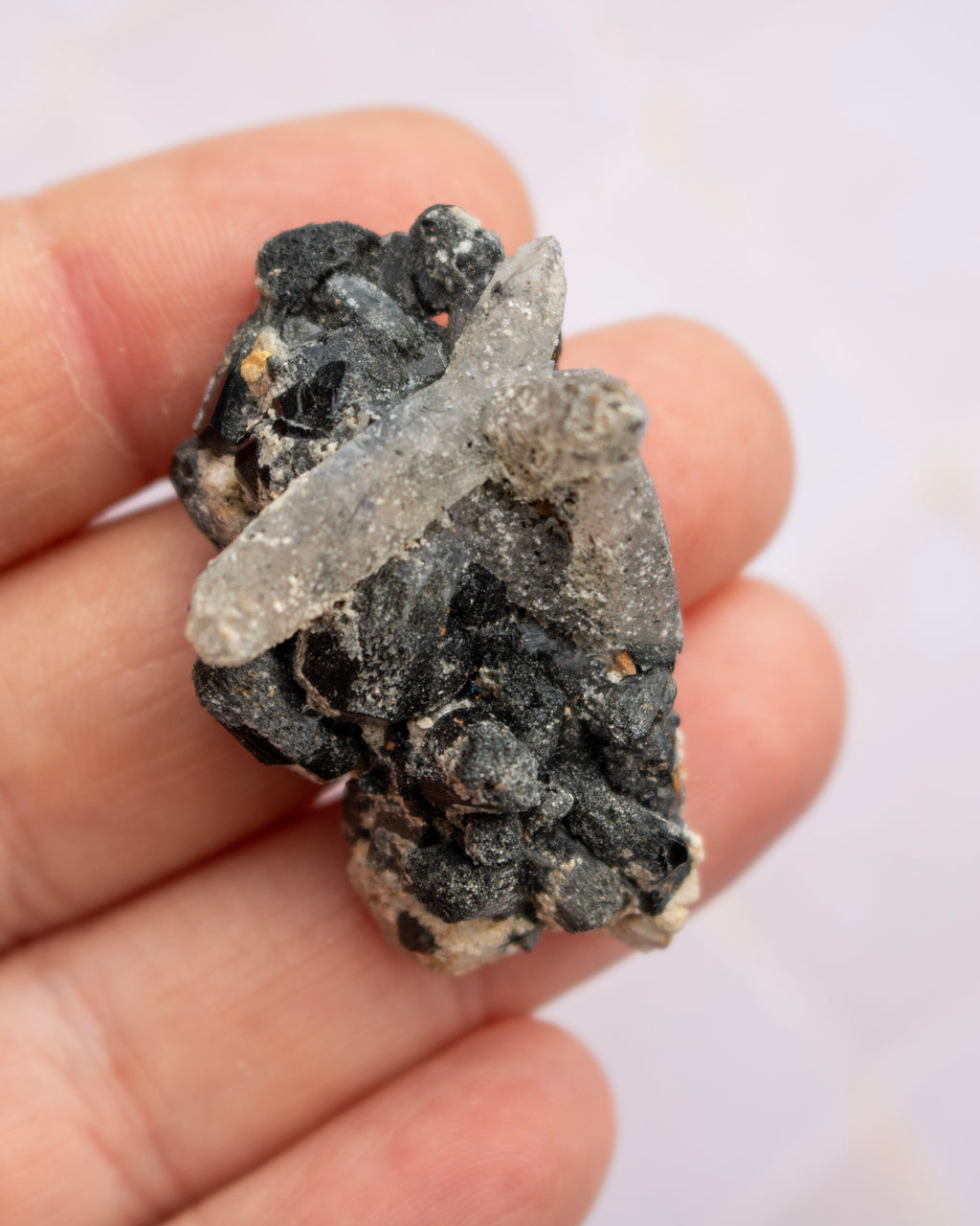 A hand holding a small mineral specimen featuring a pale translucent crystal over a dark, rough matrix.