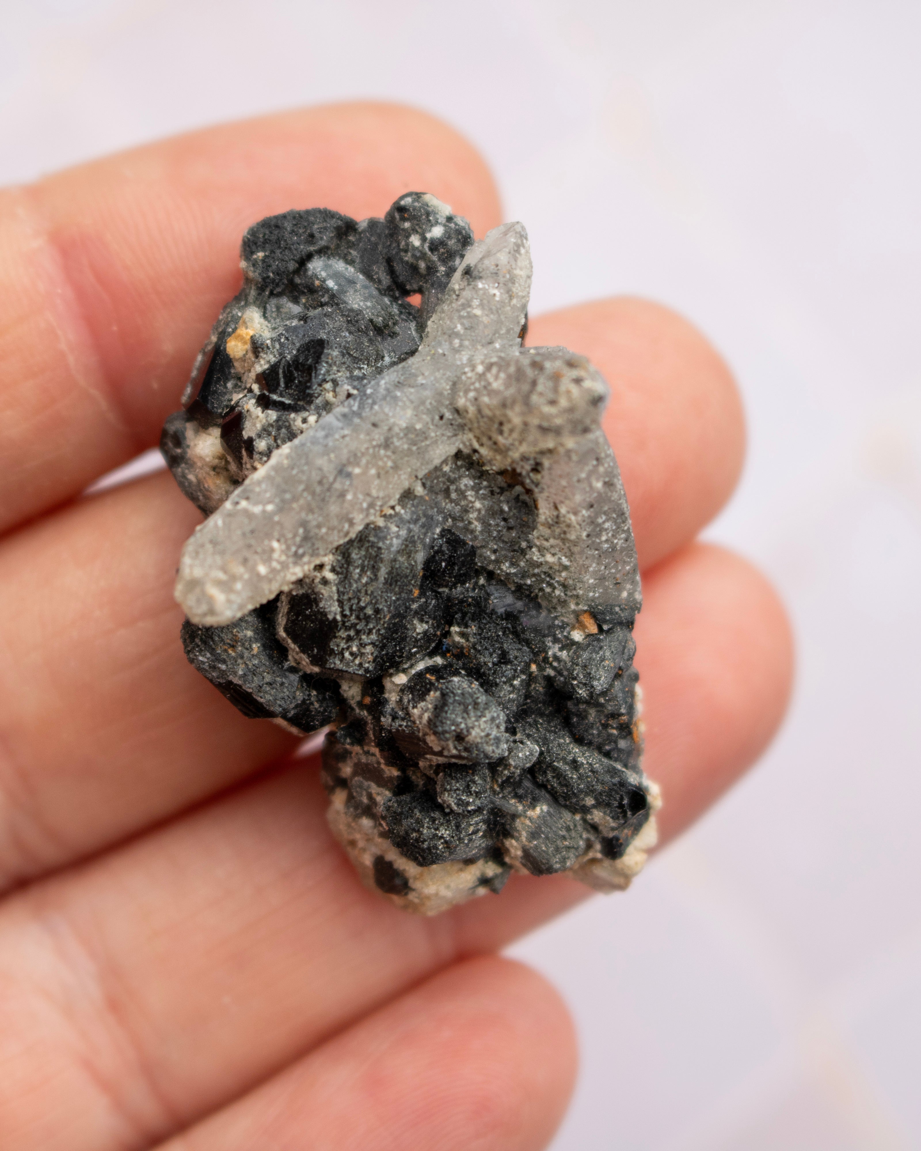A hand holding a small mineral specimen featuring a pale translucent crystal over a dark, rough matrix.