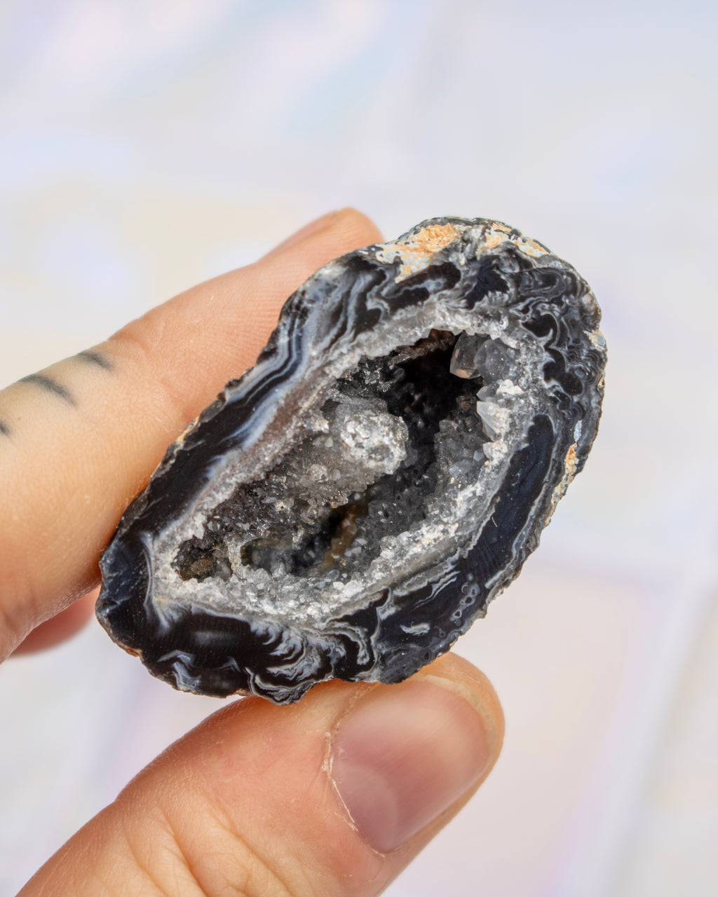 A hand holding a polished black agate geode slice with a white crystalline interior, set against a soft, out-of-focus light background.