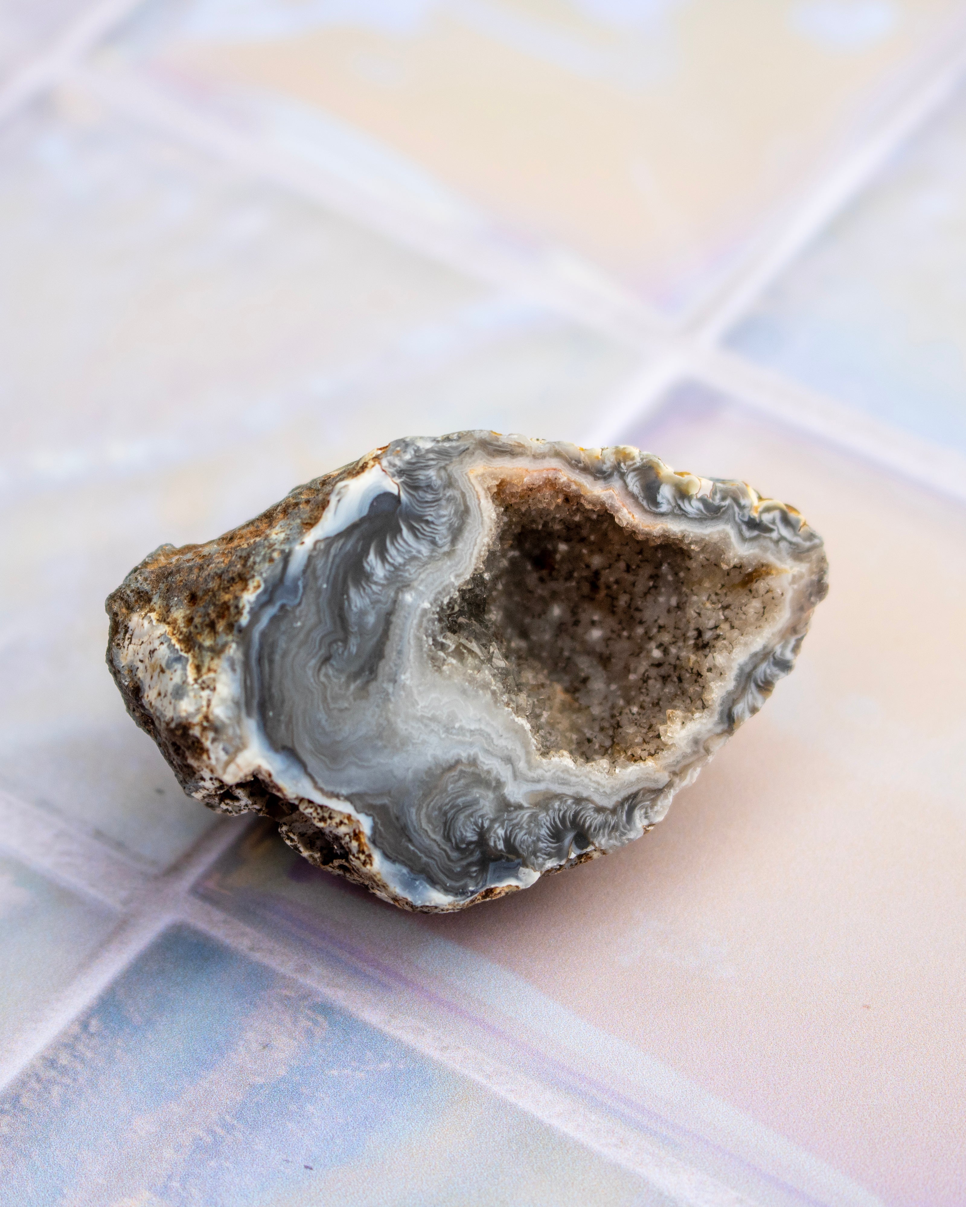 A natural geode specimen with a hollowed-out cavity, showing layered gray and white agate patterns on one side and a rough, brown crystalline interior on the other, resting on a glossy tiled surface.