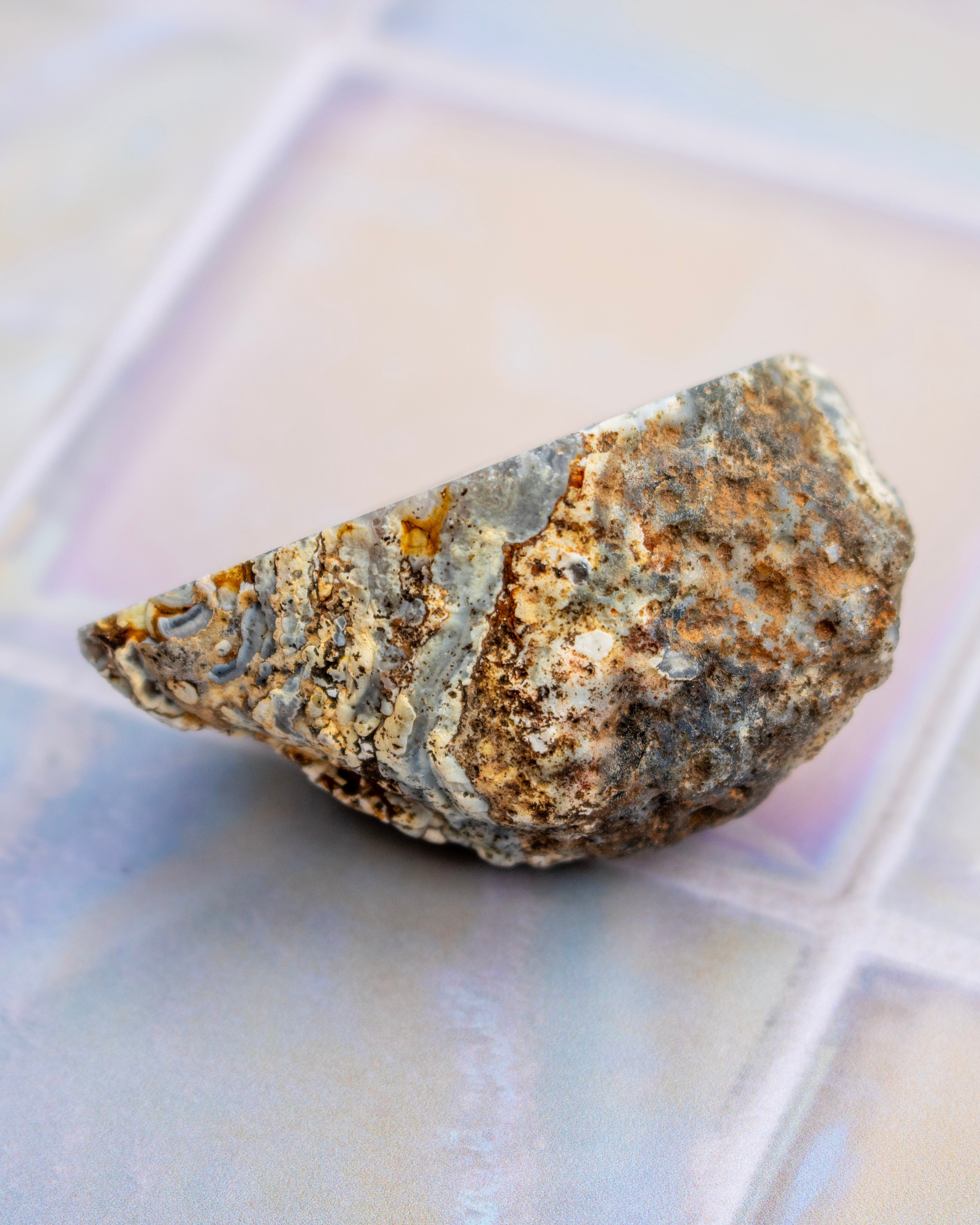 A rough, natural stone or rock specimen with a mix of gray, brown, beige, and orange tones, photographed on a softly blurred iridescent tile surface.