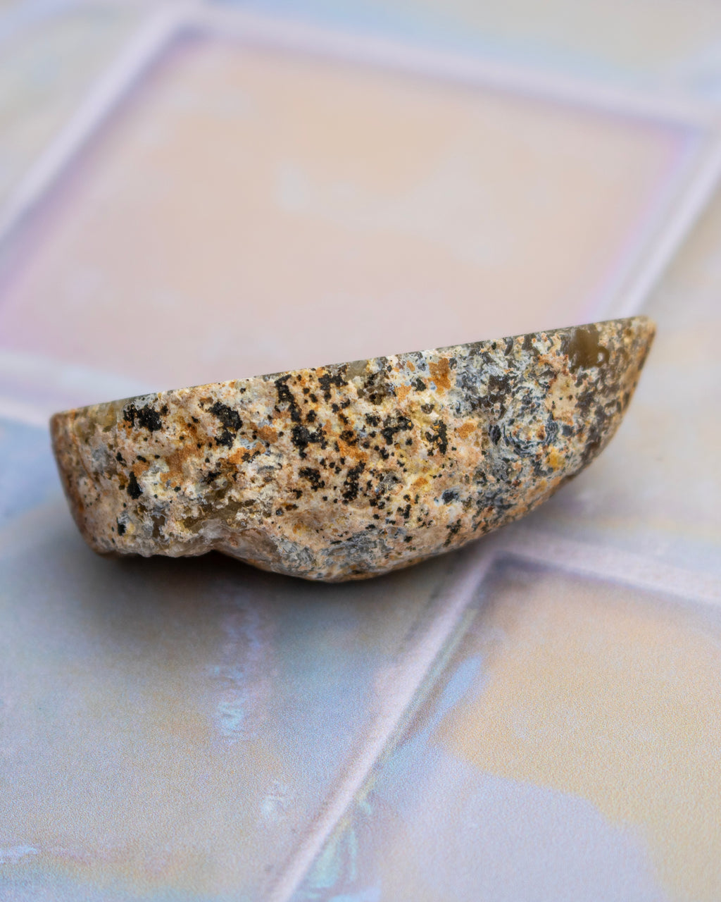 A polished wedge-shaped natural stone or quartz crystal with a speckled beige, brown, and black texture, resting on a glossy, light-colored tiled surface.
